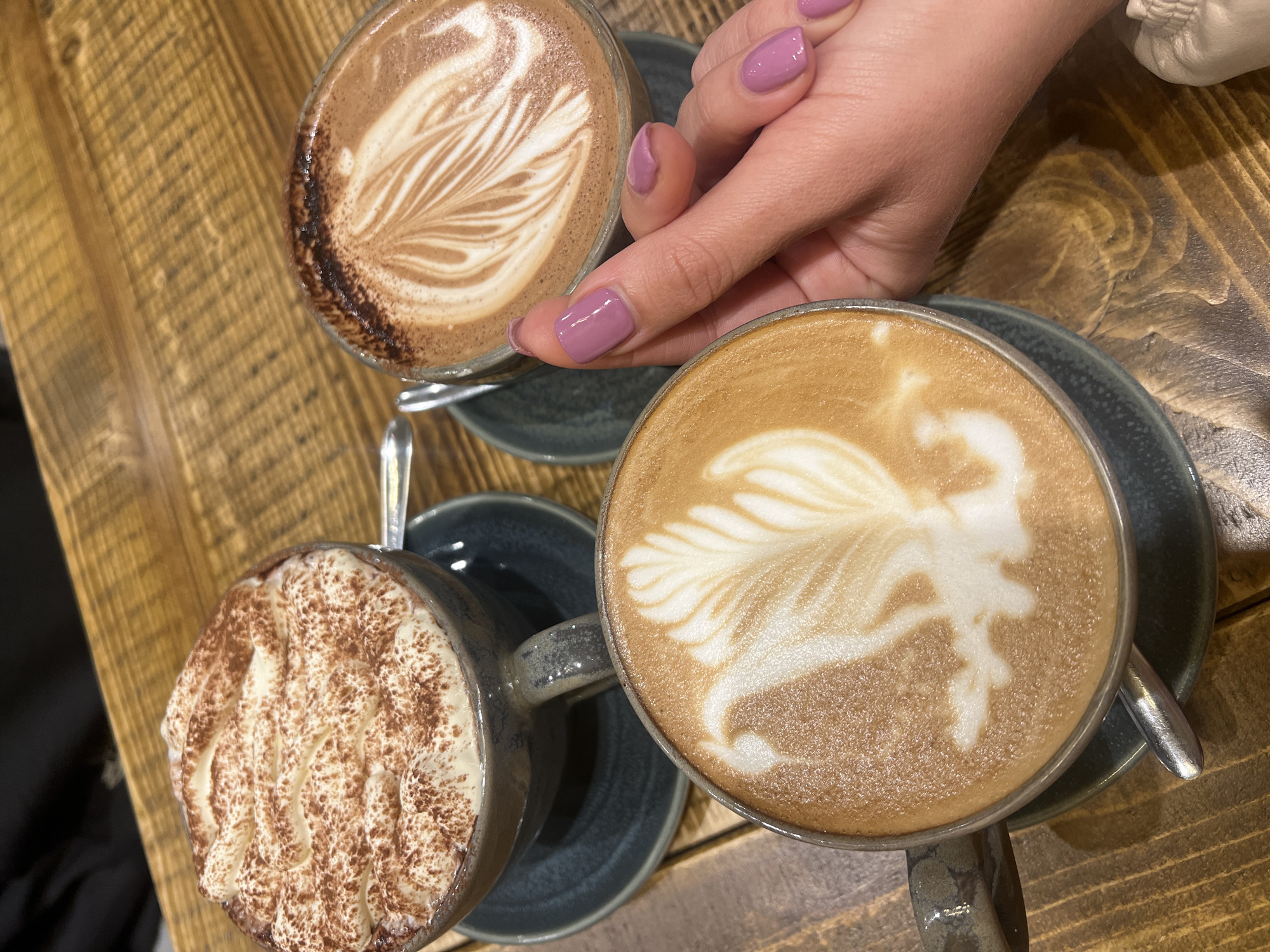 The scene is an overhead shot of a wooden table in an indoor setting, featuring three mugs of hot beverages. A person's hand with light purple nail polish is visible near one of the mugs, with their index finger slightly pointing to it. The mugs are arranged in a triangular pattern, with one containing a drink topped with cocoa powder and foamed cream, another containing latte art, and the third showing latte art in detail. Each mug sits on a dark-colored saucer. The specific location within Mole Valley, United Kingdom, is not discernible from the provided image.