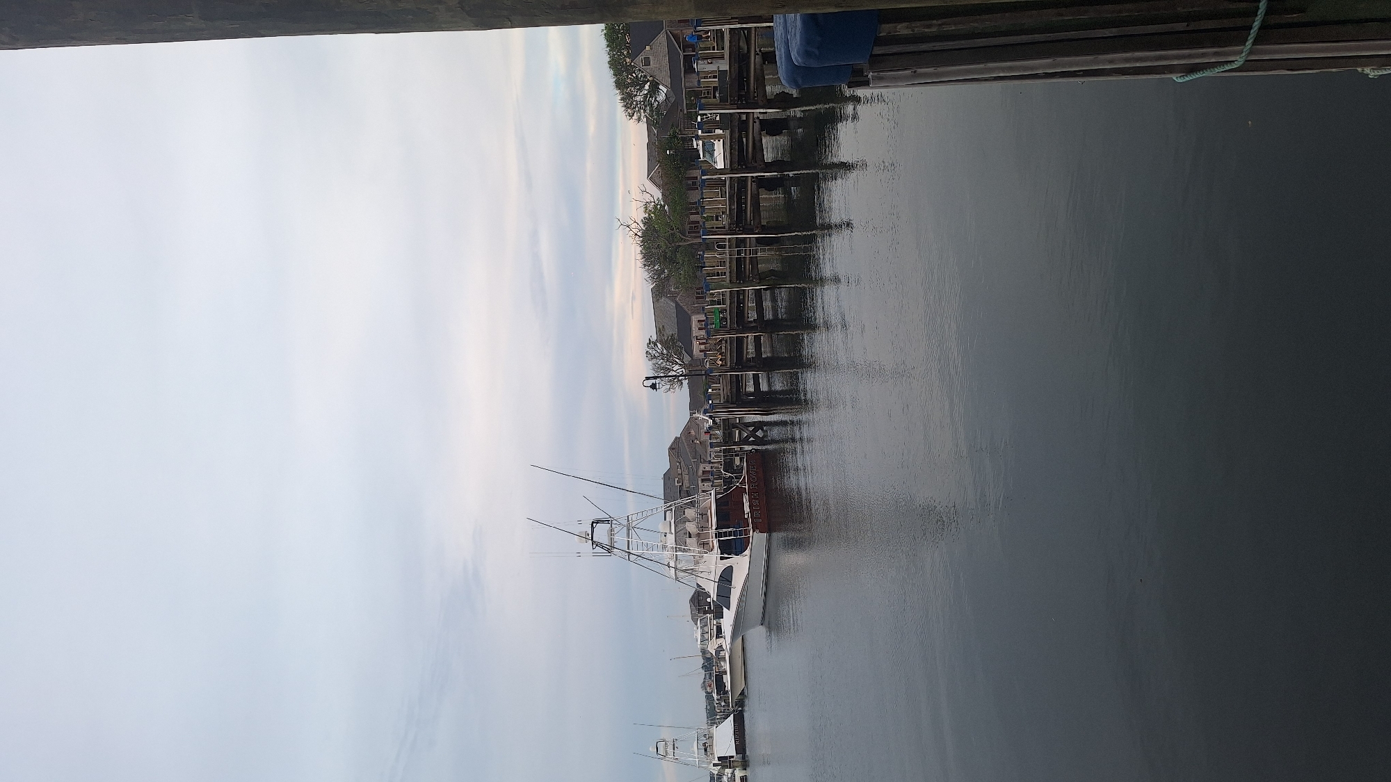 This is a photograph of a harbor scene on Nantucket, Massachusetts. Several boats are docked, including a large white sport fishing boat with the name "IRISH ROVER" visible on its hull. The boat is positioned prominently in the foreground. Beyond the boats, a wooden pier runs along the waterfront, lined with buildings that appear to be shops or restaurants. A street lamp is visible on the pier, as well as a green vehicle parked near one of the buildings. The water in the harbor is calm, reflecting the sky and the structures on the shore. The sky is overcast with soft, diffused light. A wooden piling with a green rope is in the right foreground.