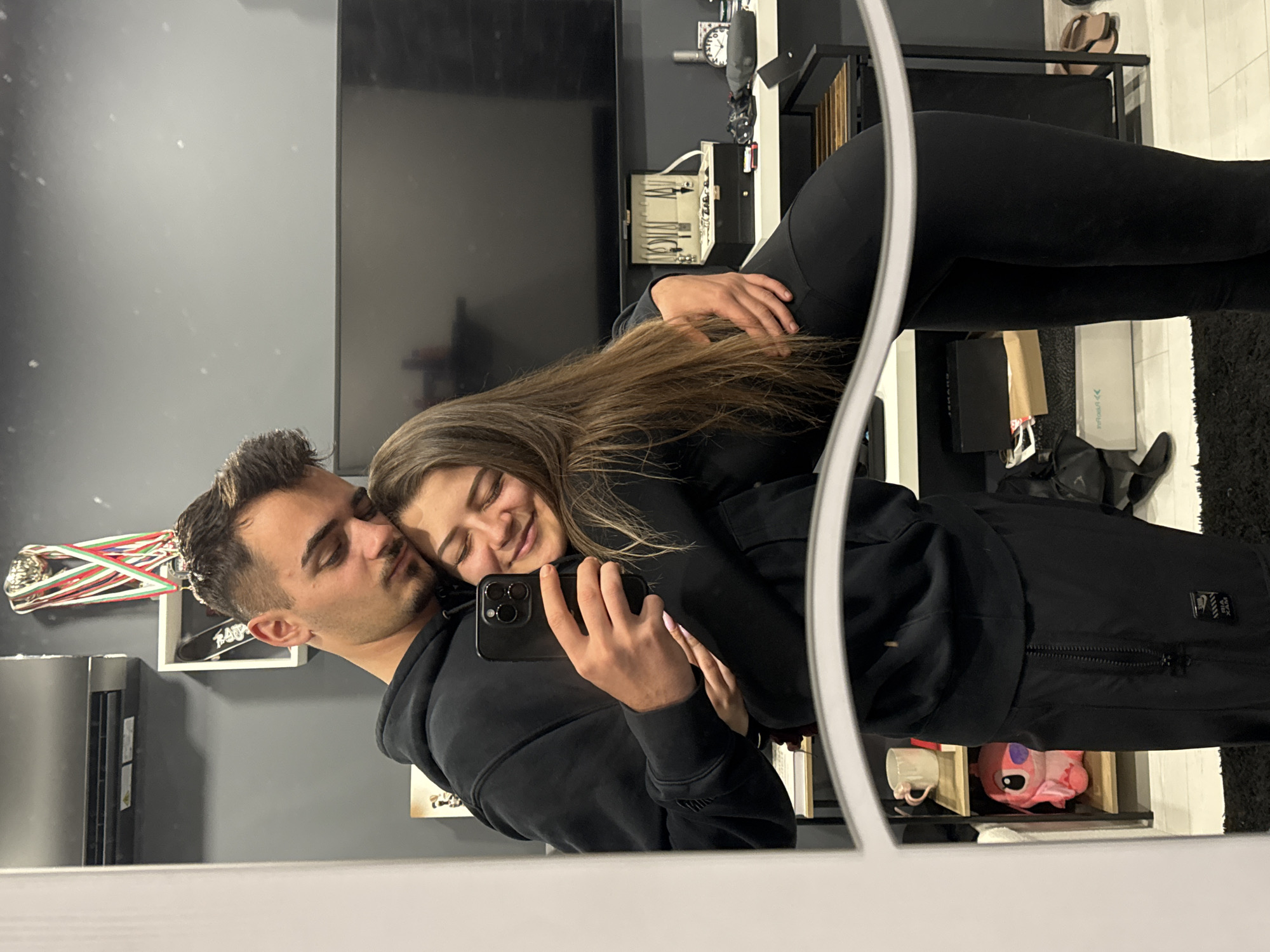 A mirror selfie captures a young couple embracing in an indoor setting. The image is taken by the woman, who holds a black smartphone in her right hand. She is leaning into the man, smiling with her eyes closed, conveying happiness and contentment. Her long brown hair flows over her shoulders. The man, with short dark hair and a beard, leans his head against hers, his eyes gently closed or looking down with an affectionate expression. Both are dressed in black casual wear; the man in a hoodie and track pants, and the woman in a hoodie and leggings.
The background, visible through the mirror, shows a contemporary interior. Behind the couple, a large black TV screen is mounted on a gray wall. To the right of the TV, a white shelf holds various items, including a white box with what appear to be dental or surgical tools, and a clock. Further to the right, a woman's leg in black leggings and a dark shoe are visible. To the left of the couple, medals hang on a wall above a white shelf. Below the man's arm, a pink plush toy (possibly Stitch) and a white mug are placed on another shelf. The floor has a black rug. The lighting suggests an indoor setting, possibly evening. No clearly legible text is visible on any objects, though there are some faint labels on clothing and boxes. The location context provided is Sofia, Bulgaria.