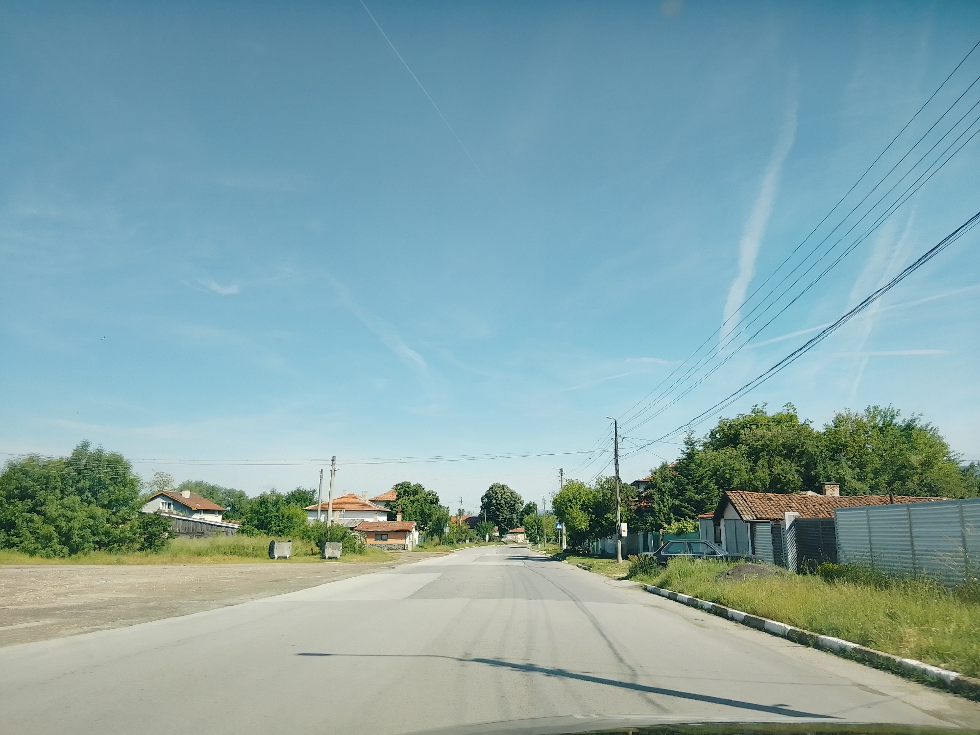 This media file captures a daytime view of a paved road in a rural or semi-rural setting, likely in Mirovo, Bulgaria. The sky is bright blue with several white contrails from aircraft visible, along with a faint, small bird in the upper left. Sunlight bathes the scene, indicating clear weather.
The asphalt road, showing signs of wear and patches, stretches straight ahead and gently curves to the right in the distance. In the foreground, a reflection from a vehicle's dashboard is visible at the bottom of the frame, suggesting the photo was taken from inside a car.
On both sides of the road, traditional-style houses with reddish-brown tiled roofs are nestled amidst abundant green foliage. These buildings are partially obscured by dense trees and shrubs, particularly on the left. On the left side of the road, there's a wide, unpaved area covered in gravel, with two rectangular grey concrete bins or containers.
The right side of the road features tall green grass and corrugated metal fences enclosing properties. A dark-colored car, possibly a sedan, is parked behind some grass and a fence on the right. Utility poles and a dense network of power lines run parallel to the road on the right, cutting across the sky. Further down the road on the right, a street light pole and a blue road sign are visible, partially hidden by trees.
No specific activity is taking place, and no people are visible. There is no discernible text within the image. The overall impression is that of a quiet village or small town road on a clear, sunny day.