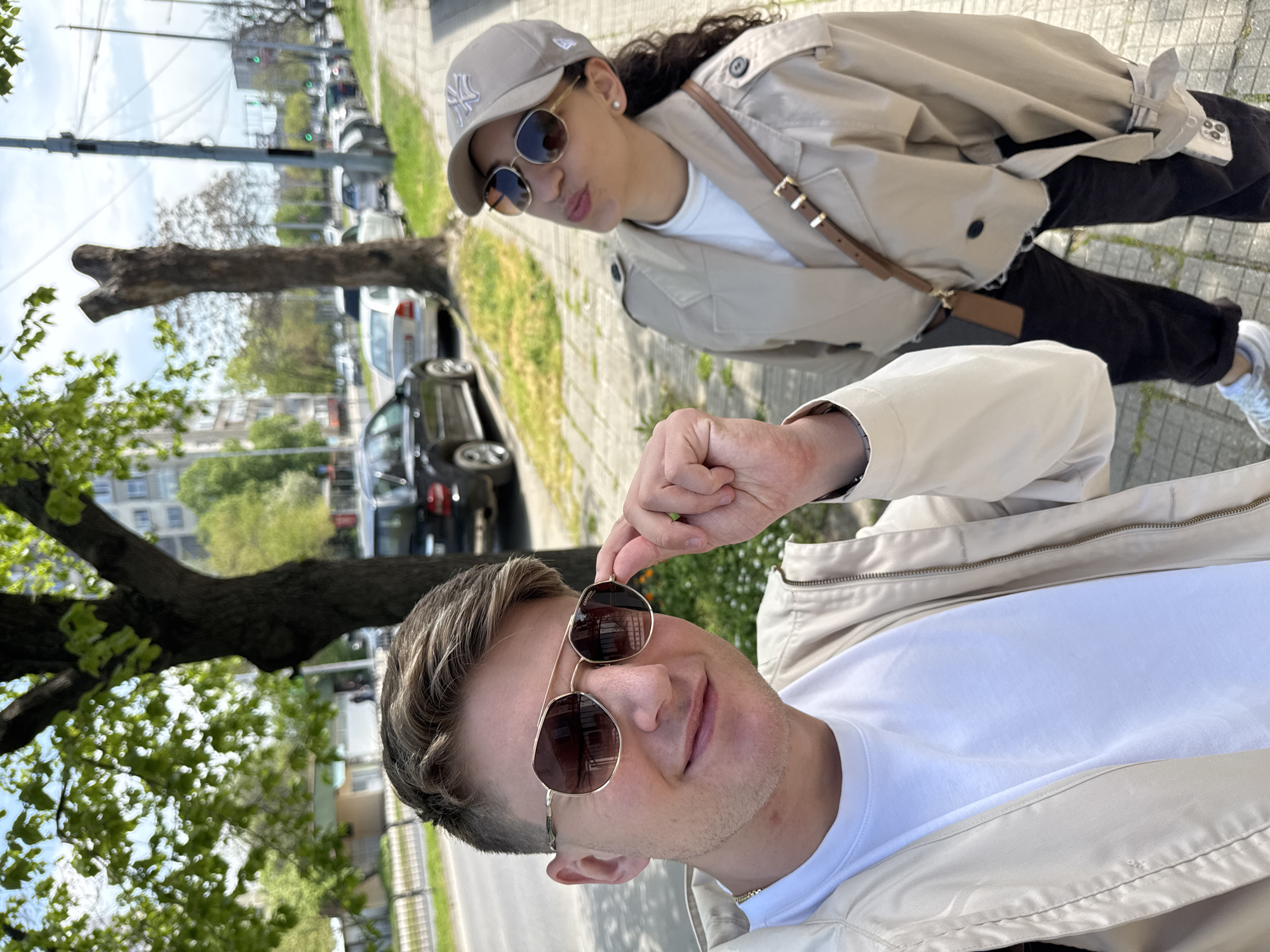 A bright, outdoor photo is presented in a portrait orientation, capturing two individuals in an urban setting, likely Plovdiv, Bulgaria. The perspective is from a low angle, with the foreground dominated by a man's upper body on the right side of the frame. He has light brown hair, light stubble, and is looking towards the viewer with a slight smile, adjusting his aviator-style sunglasses. The word "Carrera" is faintly visible on the lens. He wears a white t-shirt and a light beige jacket.
On the left side of the frame, slightly behind the man, a woman is seen from the waist up. She has dark curly hair and a subtle pouting expression as she looks towards the camera. She wears a light beige baseball cap with the New York Yankees "NY" logo, matching aviator sunglasses, a white t-shirt, and a light beige trench coat. A brown leather-strapped bag crosses her body, and an iPhone with its camera array and Apple logo is visible tucked into her trench coat pocket.
The background elements, which appear vertically oriented due to the photo's rotation, depict a sunlit street scene. Trees with green leaves line what would be the left side of the street, while numerous cars are parked along the right. A sidewalk and distant buildings complete the urban backdrop. The clear skies and strong natural light indicate a sunny day, likely late morning or early afternoon. The overall mood of the image is casual and relaxed.