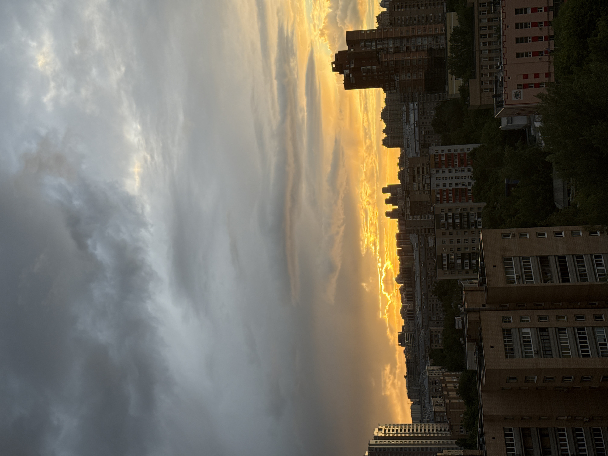 The image captures a sunset over Moscow, Russia, with a predominantly overcast sky on the left transitioning into golden hues near the horizon on the right. The sun's glow reflects off the clouds, creating a dramatic contrast. Numerous apartment buildings and high-rise structures, typical of urban Moscow, form the cityscape below the horizon. Trees are interspersed among the buildings, adding patches of green. The photograph appears to be taken from an elevated position, possibly from another tall building or a high floor. No people are visible in the shot.
