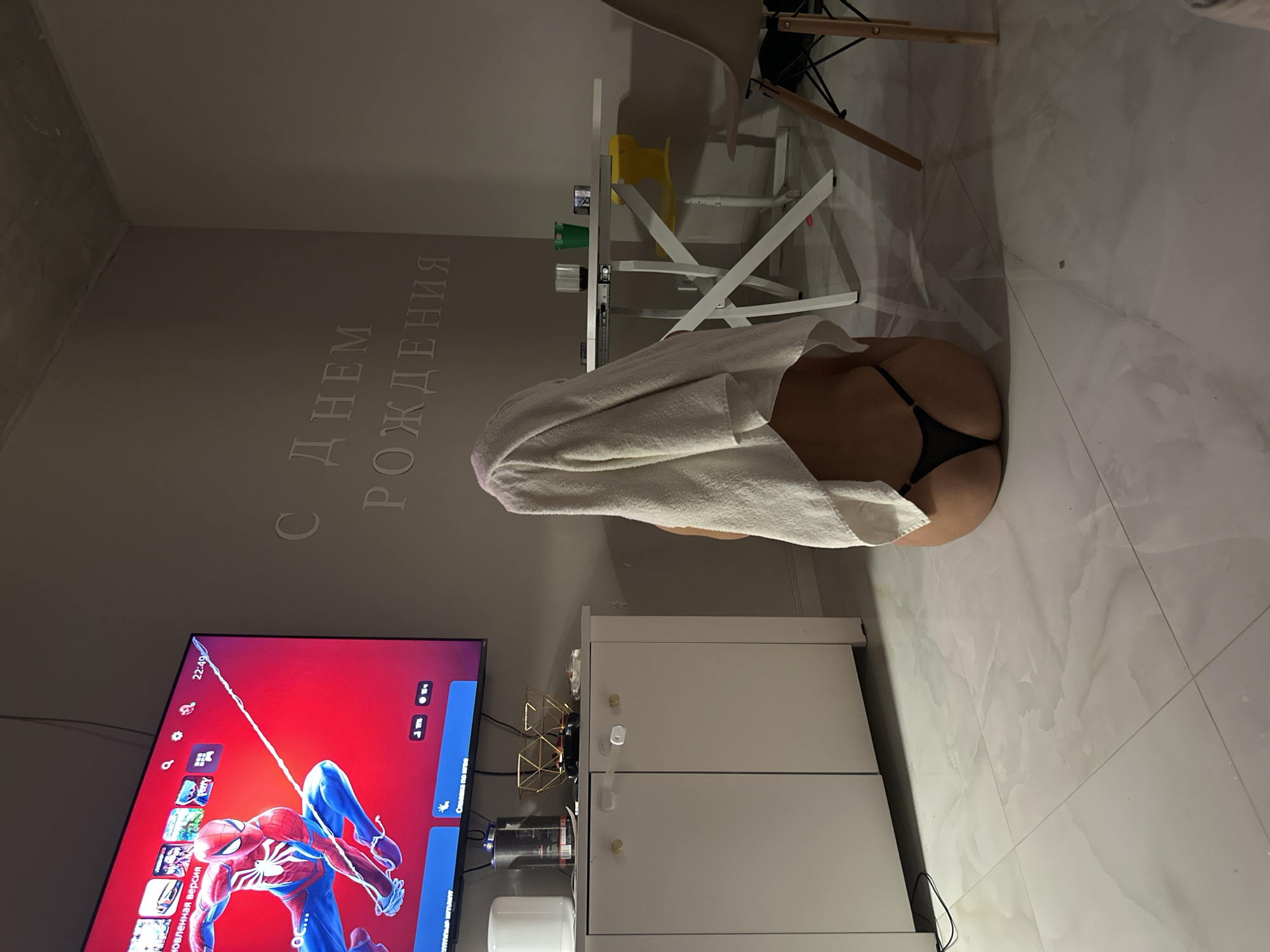 This indoor image, originally rotated 90 degrees counter-clockwise, depicts a room at night, indicated by the time 22:49 (10:49 PM) displayed on a television screen.
In the foreground, a person is kneeling or squatting on a white tiled floor, facing away from the viewer. They are wearing black thong-style underwear, and a white towel is draped over their head and back, obscuring their upper body.
To the left, a large flat-screen television is displaying a video game interface, prominently featuring the character Spider-Man in his red and blue suit. Text in Russian is visible on the screen, including "ОБНОВЛЕННАЯ ВЕРСИЯ" (Updated Version) and "Состояние сети" (Network Status). Below the TV is a white two-door cabinet, likely an entertainment unit, with a gold geometric decoration and a drink can on its surface. A white cylindrical object rests on the floor next to the cabinet.
The walls of the room are a light gray or beige. On the wall behind the person, large, carved or embossed Russian text reads "С ДНЕМ РОЖДЕНИЯ," which translates to "HAPPY BIRTHDAY." In the background, a white table holds a small yellow object and other items, and part of a chair with wooden legs is visible. The floor throughout the visible area is covered in white, subtly marbled tiles. The lighting suggests an artificial indoor light source during the evening.