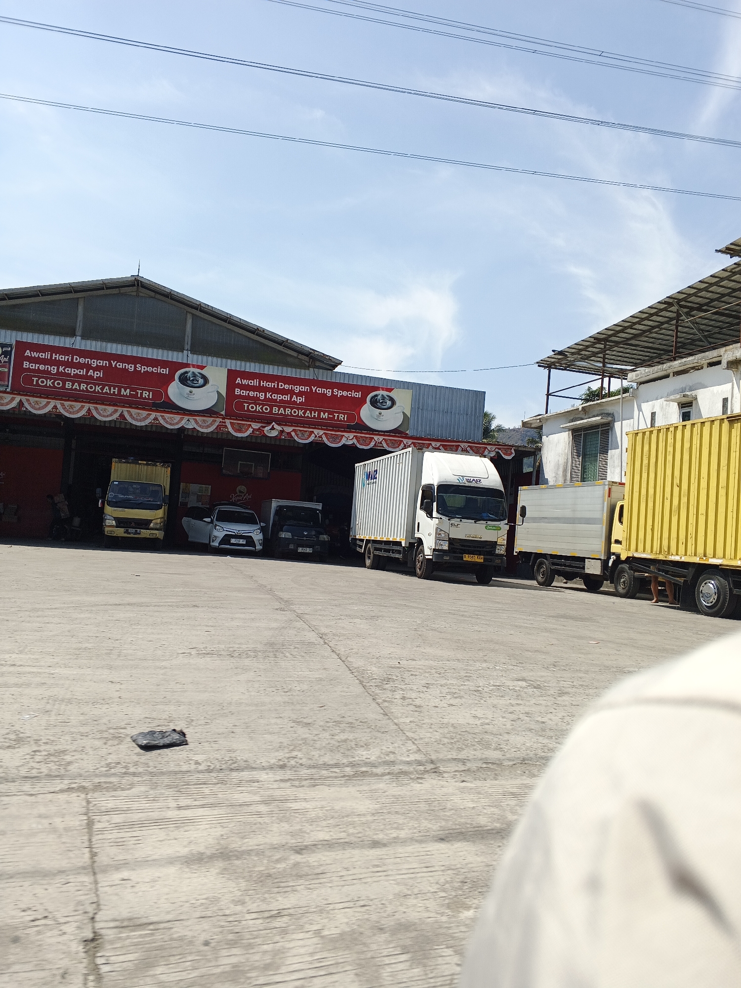 The image depicts an outdoor scene in Sukabumi, Indonesia, likely a commercial area or depot. In the foreground, a concrete surface is visible, with a dark, irregular object lying on it. A portion of a person's shoulder and head, covered in light-colored fabric, is also in the lower right corner.
The midground features several vehicles parked on the concrete. From left to right, there is a yellow truck, a white car, and a black truck. Behind these, a white truck with a cargo container is prominent, followed by another truck with a white cargo container and a yellow cargo container truck. The trucks appear to be parked in a line.
The background reveals the facade of a building with a red exterior. A banner with Indonesian text, including "Awali Hari Dengan Yang Special Bareng Kapal Api TOKO BAROKAH M-TRI," is displayed prominently across the top of the building's opening. Above the building, power lines stretch across a bright blue sky with some scattered clouds. To the right of the main building, there is another structure with a corrugated metal roof supported by a metal frame. The overall environment suggests a logistics or distribution point.