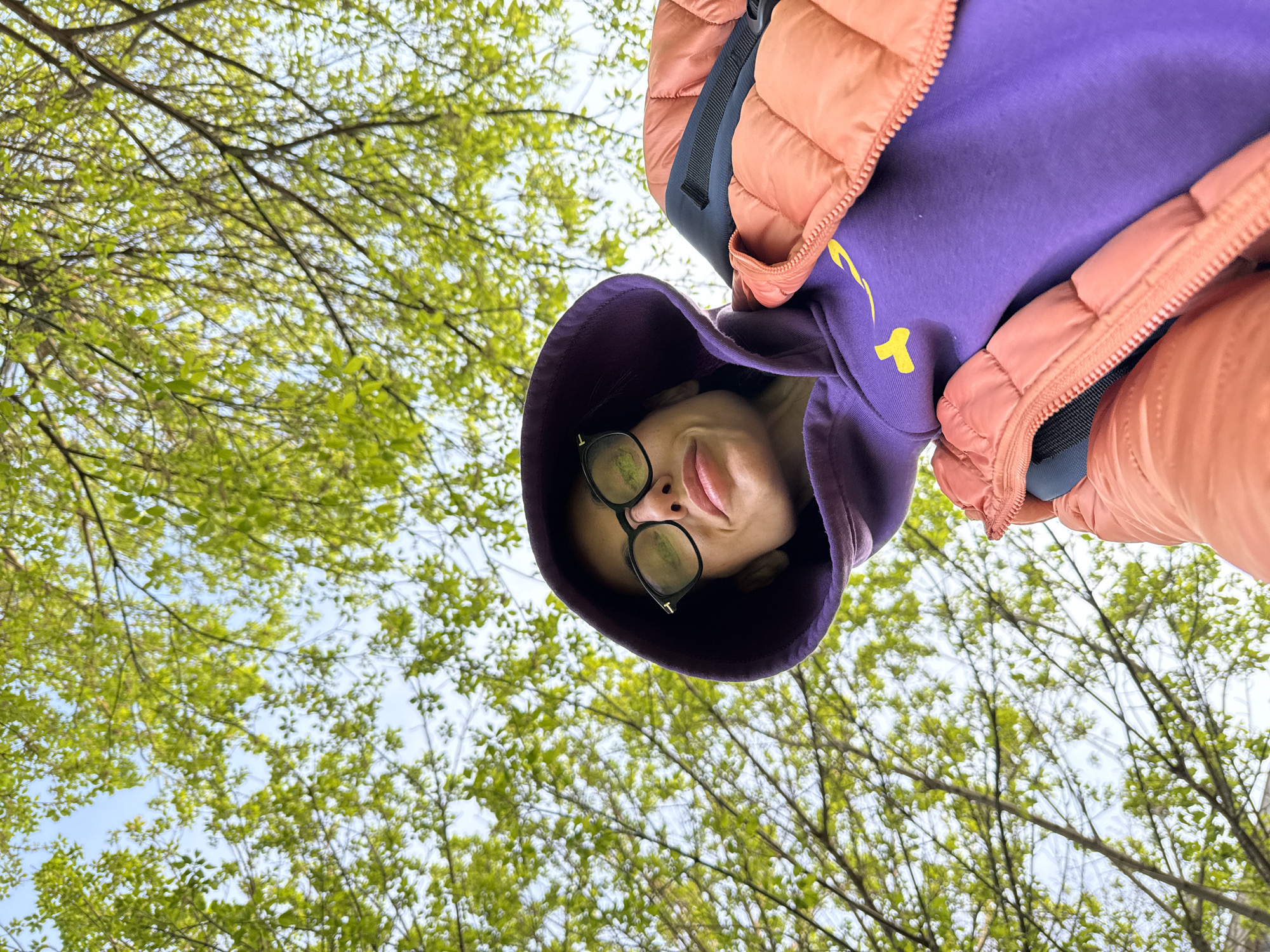 A low-angle view of a person wearing black eyeglasses and a purple hooded sweatshirt under a peach-colored jacket. The person appears to be looking down at the camera. Behind them, a blurred view of trees with green leaves against a bright sky. No specific identifying details indicate the location is Pleven, Bulgaria.