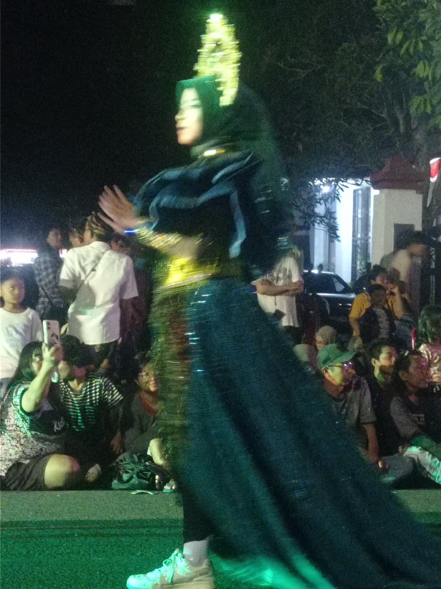 A dancer in a dark blue dress and a gold headpiece is the central figure in this nighttime outdoor scene in Pandean, Indonesia. The dancer, facing to the right, has her hands raised. She is performing on a green surface, possibly a stage or illuminated ground. A crowd is gathered behind her, some people are seated and others are standing. Several individuals in the crowd are looking towards the dancer, and one person in the foreground is holding up a phone, likely taking a picture or recording a video. The background includes trees, a building with windows, and a car. The overall lighting is dim, with green and yellow lights illuminating the scene.