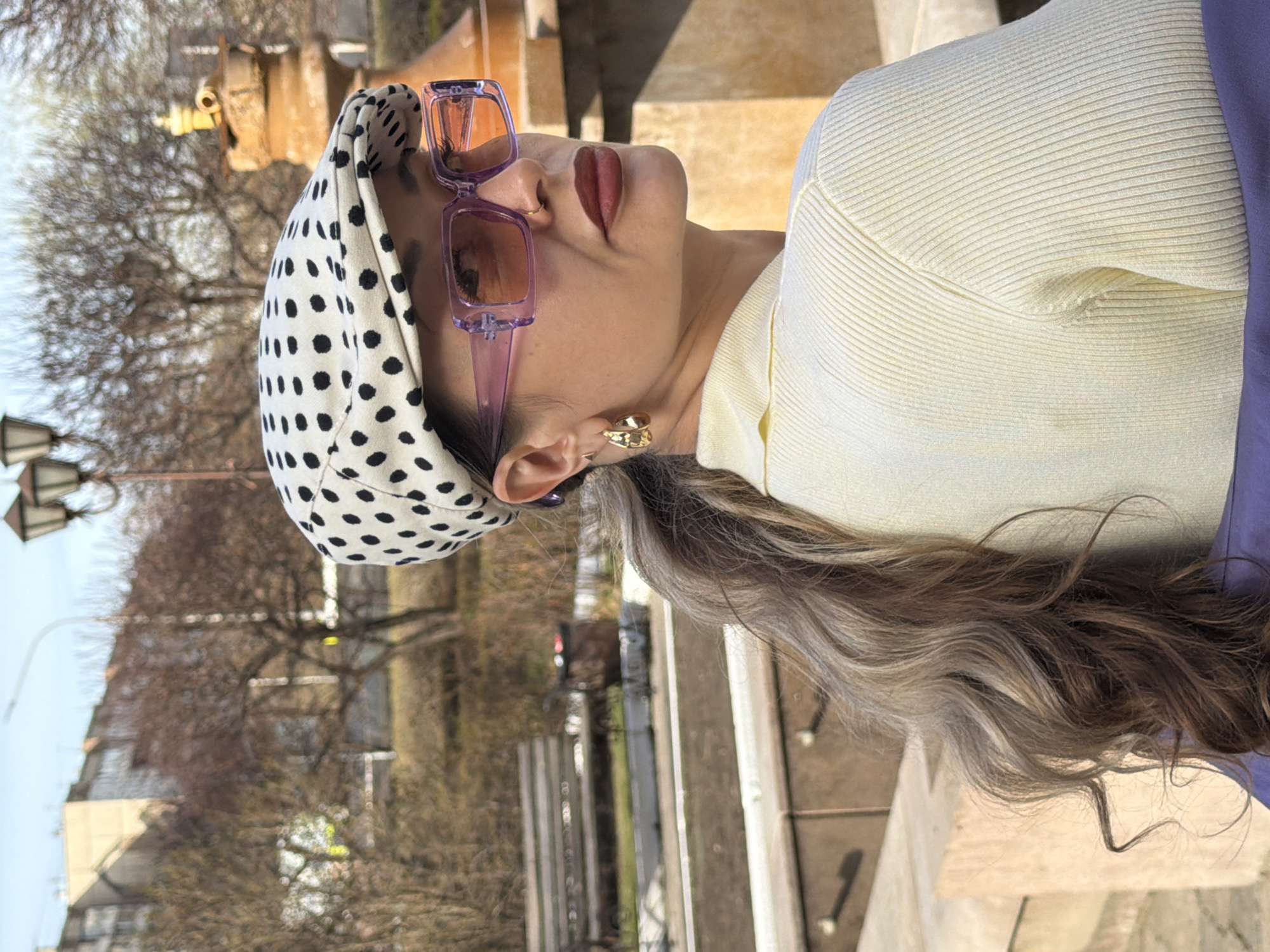 An outdoor portrait photograph shows a person, likely a woman, captured from the mid-chest up in a profile view, looking upwards and to her left with a slight head tilt. She is wearing a white headscarf with black polka dots and purple-framed sunglasses with tinted lenses perched on her nose. Her lips are painted a dark red or maroon color, and she wears a gold-toned, abstractly-shaped earring in her right ear. She is dressed in a light yellow, ribbed long-sleeved top, with a portion of a purple garment visible on her left shoulder. Her long, wavy hair, featuring a mix of dark brown and lighter, possibly ash-blonde or gray, strands, flows behind her.
The background, set in an urban or park environment in Krasnoyarsk, Russia, reveals bare trees with detailed branches, suggesting late winter or early spring. Buildings and a dark street lamp are partially visible in the blurred distance. The scene is illuminated by natural daylight, indicating clear weather. The person appears to be posing for the photo, conveying a composed and stylish demeanor. No text is visible in the image.