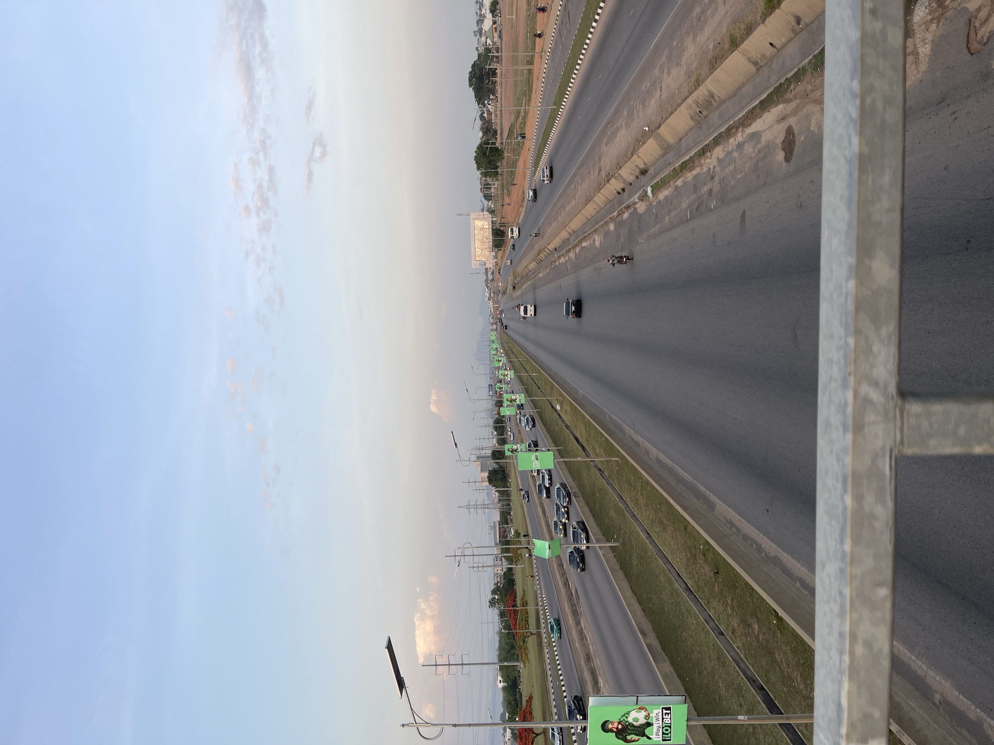The image depicts a multi-lane highway in Galadimawa Village, Nigeria, viewed from an elevated position, likely a pedestrian overpass. The road stretches into the distance, with light vehicular traffic visible. Green medians separate the lanes. Advertisement banners are spaced along one side of the highway. The sky is light blue to the left, transitioning to a dusky orange-grey towards the horizon. Several utility poles and wires run parallel to the highway on one side. The foreground includes a metal railing, presumably part of the overpass structure. The landscape beyond the road appears to be a mix of trees and low buildings.