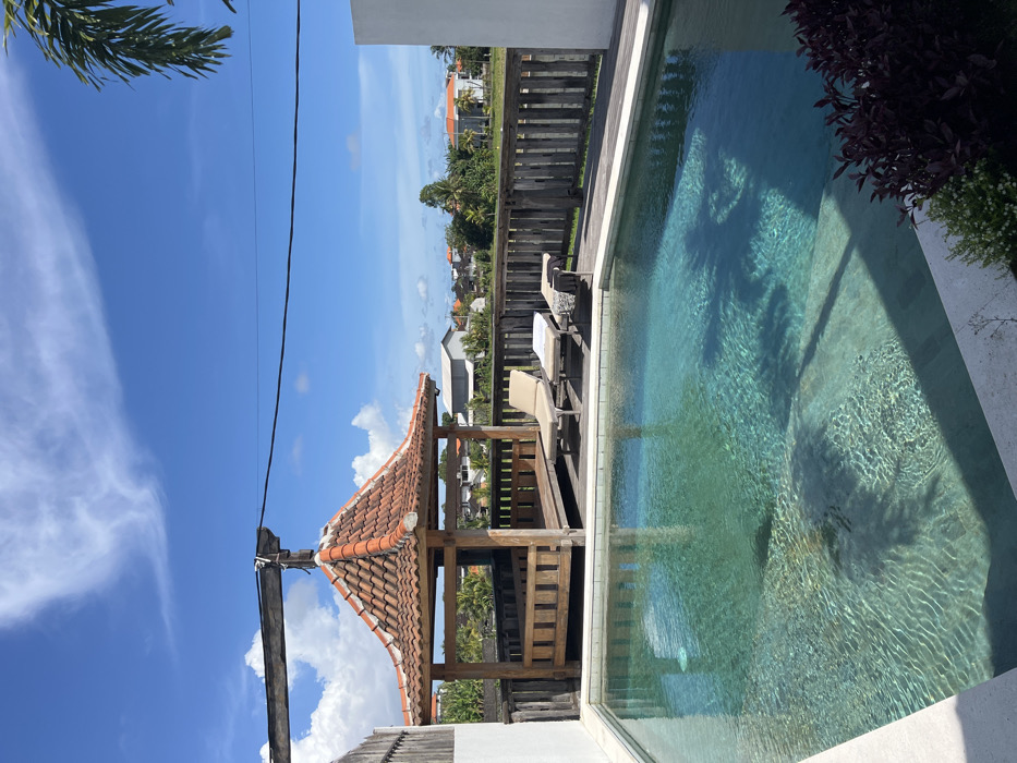 The image depicts an outdoor leisure area in Belalang, Indonesia, under a clear blue sky with scattered white clouds. In the foreground, a rectangular swimming pool with turquoise-blue water occupies the lower left and central sections. The pool bottom is visible, showing sun reflections and faint shadows. Light-colored coping surrounds the pool. To the rear of the pool, a wooden deck extends across the midground.
On this deck, a hexagonal or octagonal wooden gazebo with a reddish-brown tiled hip roof stands prominently in the center-rear. Its sides feature open wooden balustrades. To the right of the gazebo, two beige lounge chairs are positioned on the wooden deck. The chair closer to the gazebo is empty. The second chair has a white towel draped over its headrest and a dark towel at its foot.
A weathered wooden picket fence runs along the edge of the deck, forming a barrier across the midground. Beyond the fence, the background reveals a mix of green vegetation, possibly fields, and scattered residential or commercial buildings with light-colored walls and reddish-brown roofs. Several palm trees are visible among the distant structures and greenery. A thick, weathered wooden beam is visible in the upper left, with a thin black cable stretching horizontally across the upper midground. A palm frond is partially visible in the upper right corner. No individuals are present in the scene.