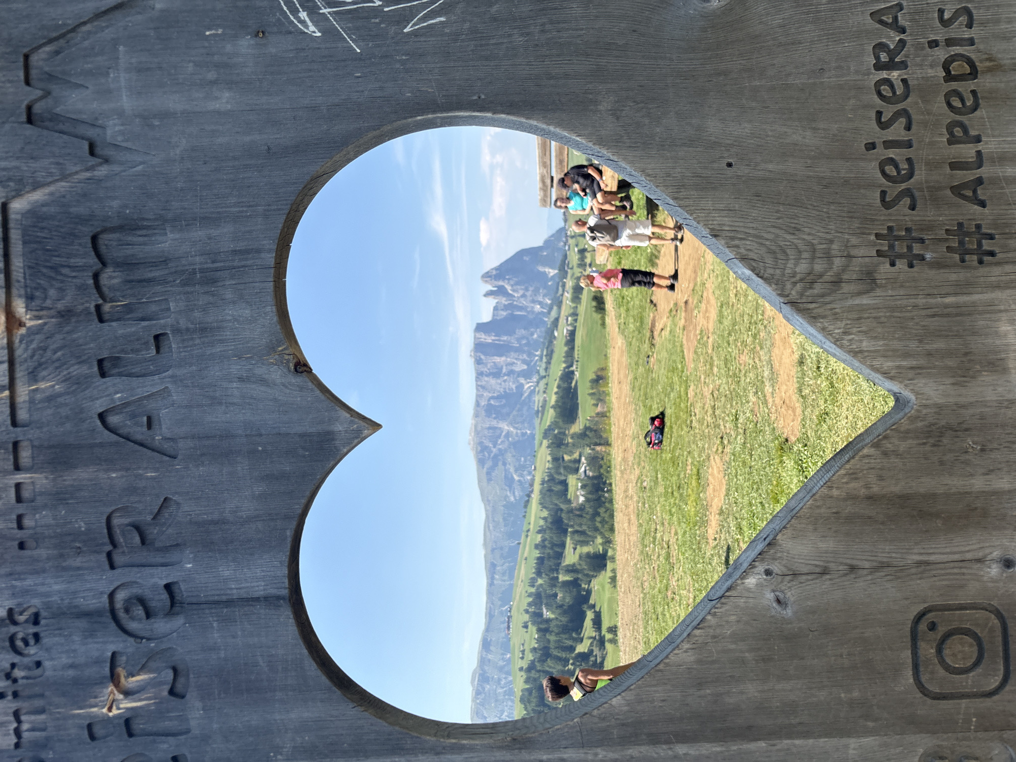 The image shows a wooden frame carved into the shape of a heart, with a scenic mountain landscape visible through the opening. The wooden frame has carved text that appears to say "mrites" and "iser ALM". On the lower right of the frame, there are two hashtags carved into the wood: "#seisera" and "#Alpebis". In the foreground, within the heart frame, a woman is standing and looking out at the landscape. Further into the landscape, a group of people are gathered, with some sitting on a bench and others standing. The landscape itself consists of rolling green hills dotted with trees, and in the background, majestic, jagged mountains rise against a clear blue sky. The lighting suggests it is a sunny day. The location is identified as the Alpe di Siusi (Seiser Alm) in Kastelruth - Castelrotto, Italy, a popular area known for its stunning alpine scenery.