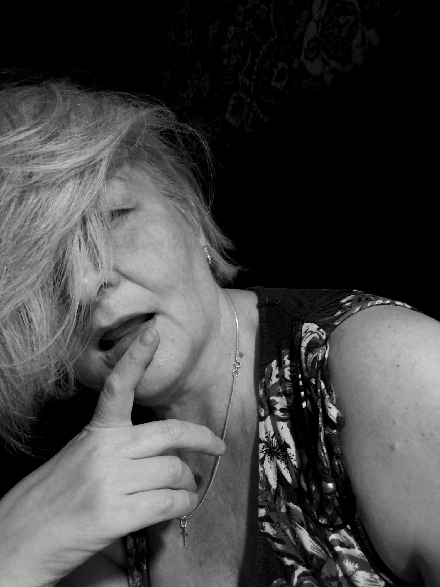 This is a black and white, close-up, indoor portrait of an older woman. Her face is partially obscured by her blonde hair, which is styled in a windswept manner. She has her eyes closed and her mouth is slightly open, with a finger touching her lips, suggesting a moment of introspection or sensuality. She is wearing a patterned top with floral motifs and a delicate silver necklace with a small cross pendant. The background is dark and textured, possibly a fabric or wallpaper, with a hint of a floral pattern visible in the upper right corner. The lighting is dramatic, casting shadows and highlighting the woman's features. The overall mood is intimate and contemplative. The location is specified as Krasnodar, Russia. There is no visible text in the image.