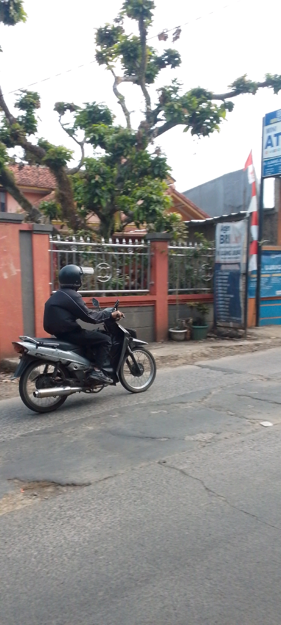 In the city of Cilaku, Indonesia, a person wearing a black jacket and helmet is riding a silver motorcycle on a paved road. The motorcycle has black handlebars and a silver exhaust pipe. The rider is facing left and is holding the handlebars. The road surface is gray and cracked, with some patches of damage. In the background, there is a red wall and a metal fence. Beyond the fence, there are trees and a building with a red roof. To the right, there is a blue sign with white lettering, and a red and white flag. The sky is overcast.