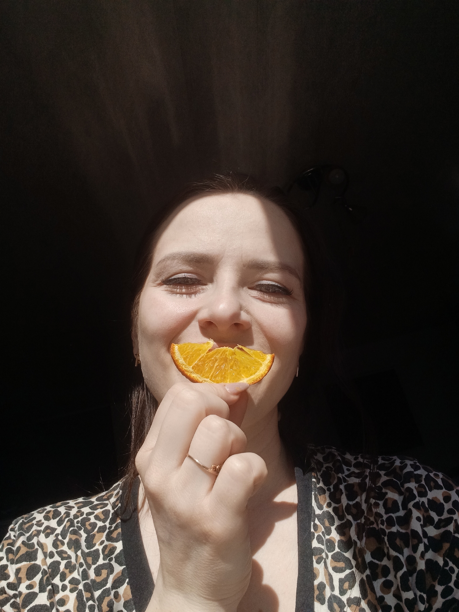 A portrait-style photograph captures a smiling woman playfully holding a slice of orange over her mouth, partially obscuring it. She has dark hair, which frames her face, and dark eyes that crinkle at the corners with her joyful expression. Strong, direct light, likely natural sunlight, illuminates her face from the front-left, creating high contrast and casting noticeable shadows, particularly on her forehead and under her chin. Her overall demeanor appears cheerful and happy.
She is wearing a top featuring a distinctive brown and black leopard print pattern, with a solid black trim visible on the shoulder area. A simple gold-toned ring adorns the ring finger of her right hand, which is holding the vibrant orange slice. The background is predominantly dark and indistinct, suggesting an indoor setting with minimal ambient light beyond the direct source illuminating the subject. In the upper right portion of the dark background, a faint outline, possibly belonging to a pair of dark-framed glasses, can be vaguely discerned. There is no visible text in the image. The strong lighting indicates the photograph was taken during the daytime. This image was captured in Rusalevka, Russia.