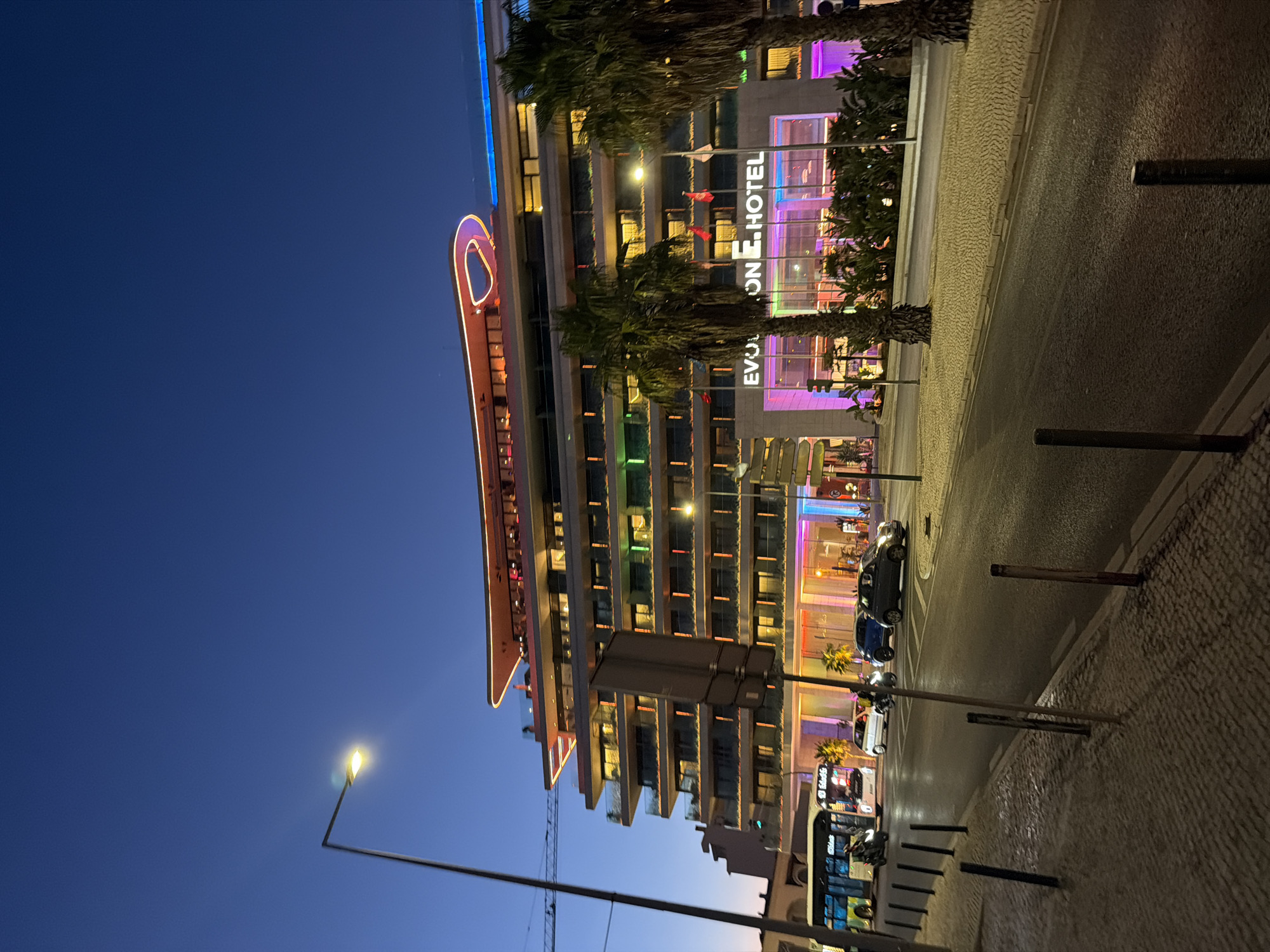 The image depicts the EVOLUTION HOTEL in Cascais, Portugal, at twilight. The modern hotel building features multiple levels with balconies and is illuminated by colorful lights, including green, purple, and orange. The top of the building has a distinctive curved structure with neon lighting. In front of the hotel, palm trees are visible, along with flags. A street runs in front of the hotel, with several cars and motorcycles on it. A bus is also seen on the left side of the image. The sidewalk on the left is paved with cobblestones, and a streetlamp is visible. The sky is a deep blue, indicating either early evening or late twilight.