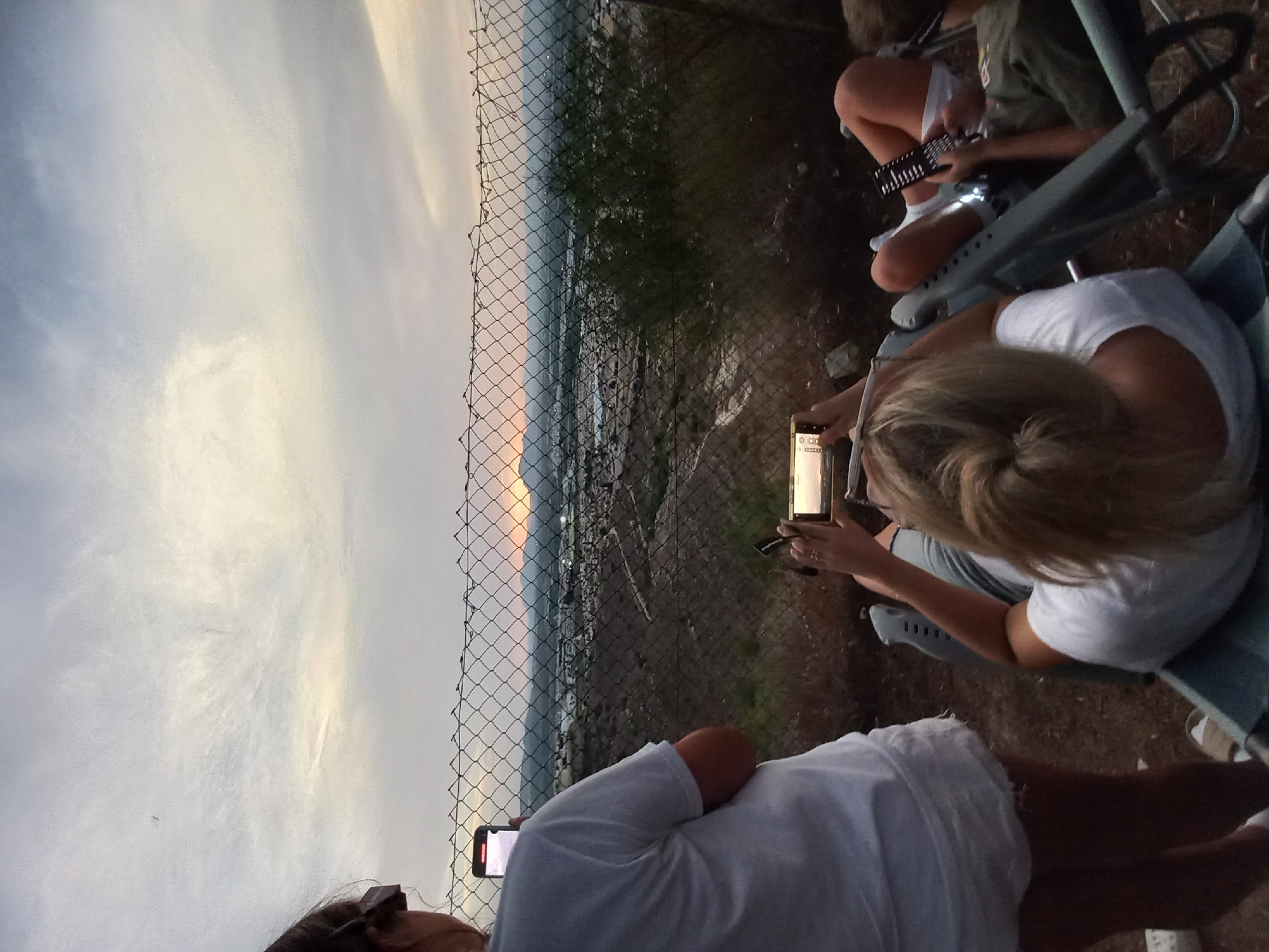 An outdoor, high-angle, medium shot shows three individuals, two women and one person of indeterminate gender, viewed from behind as they observe a sunset over Urbanización Ciudad Quesada, Spain. The foreground is occupied by the backs of their heads and upper bodies, with one woman standing and two sitting in lounge chairs. The standing woman, on the left, wears a white t-shirt and white shorts, holding a smartphone vertically. The two seated individuals are on the right. The woman in the center, with blonde hair tied back, wears a white t-shirt and is holding a smartphone horizontally. The person to her right, partially visible, wears a green shirt and white socks, holding a rectangular object, possibly a remote control, in their right hand. All three are positioned behind a chain-link fence that runs horizontally across the mid-ground. Beyond the fence, a city sprawls across a valley, with buildings and lights visible. The setting sun casts a warm orange and yellow glow on the horizon, framed by mountains in the distance. The sky is overcast with wispy clouds, transitioning from a bright, lighter hue near the horizon to a darker, grayish-blue towards the top of the frame. The ground in the foreground appears to be dirt and gravel, with some sparse vegetation.