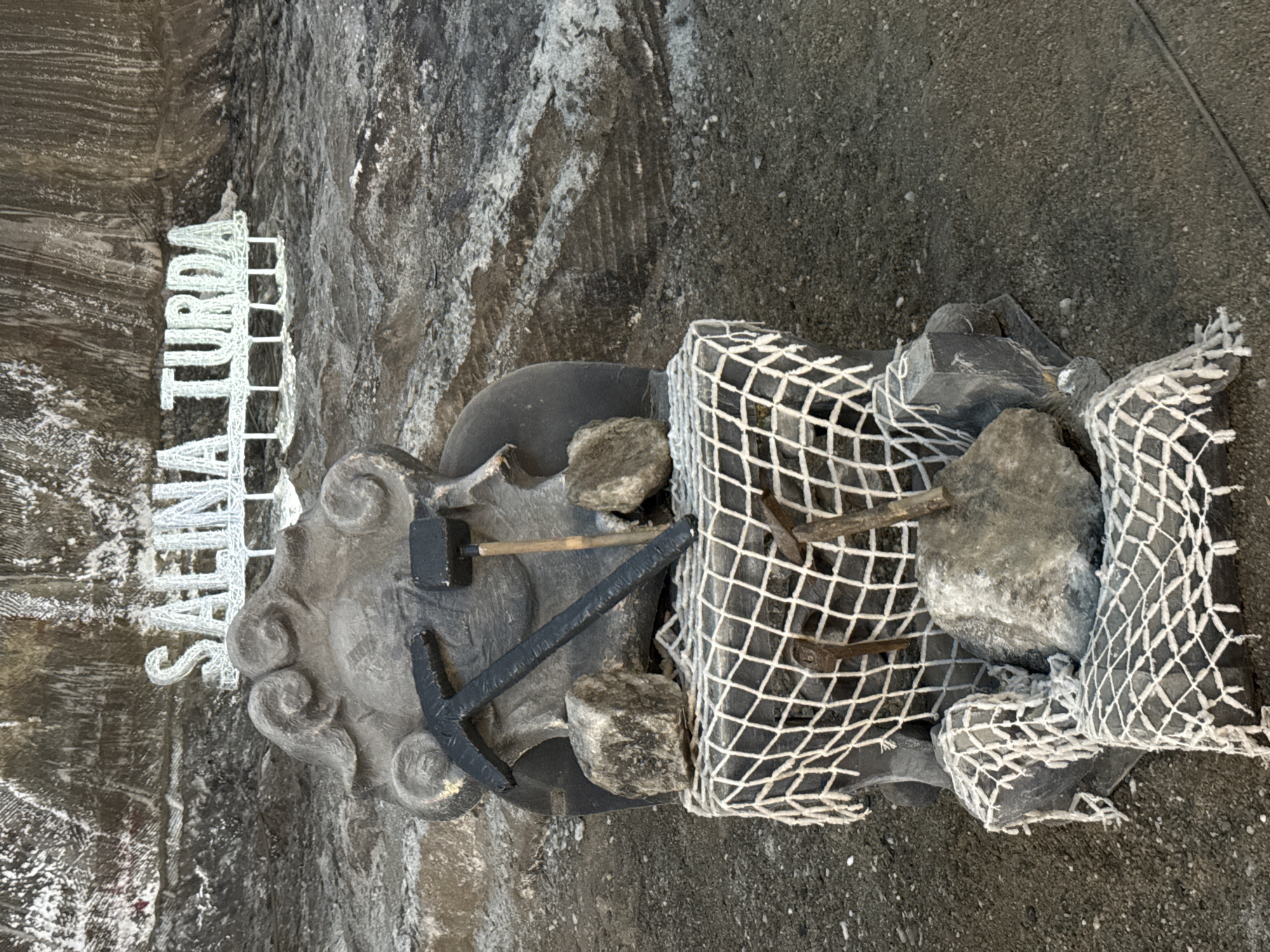 In the city of Craiova, Romania, within the Salina Turda salt mine, a monument stands amidst the rough, textured salt walls and floor. The monument is a carved, dark grey stone representation of a seated figure, possibly a miner, with decorative swirls on its headpiece. Two mining tools, a pickaxe and a hammer, are placed prominently on the figure's lap. The monument is adorned with a white net draped over its lower portion, with several large, rough-hewn rocks placed around and on top of the net. Two smaller, rusty metal tools are also partially visible sticking out from the net. The overall scene is one of remembrance and historical representation within the unique subterranean environment of the salt mine.