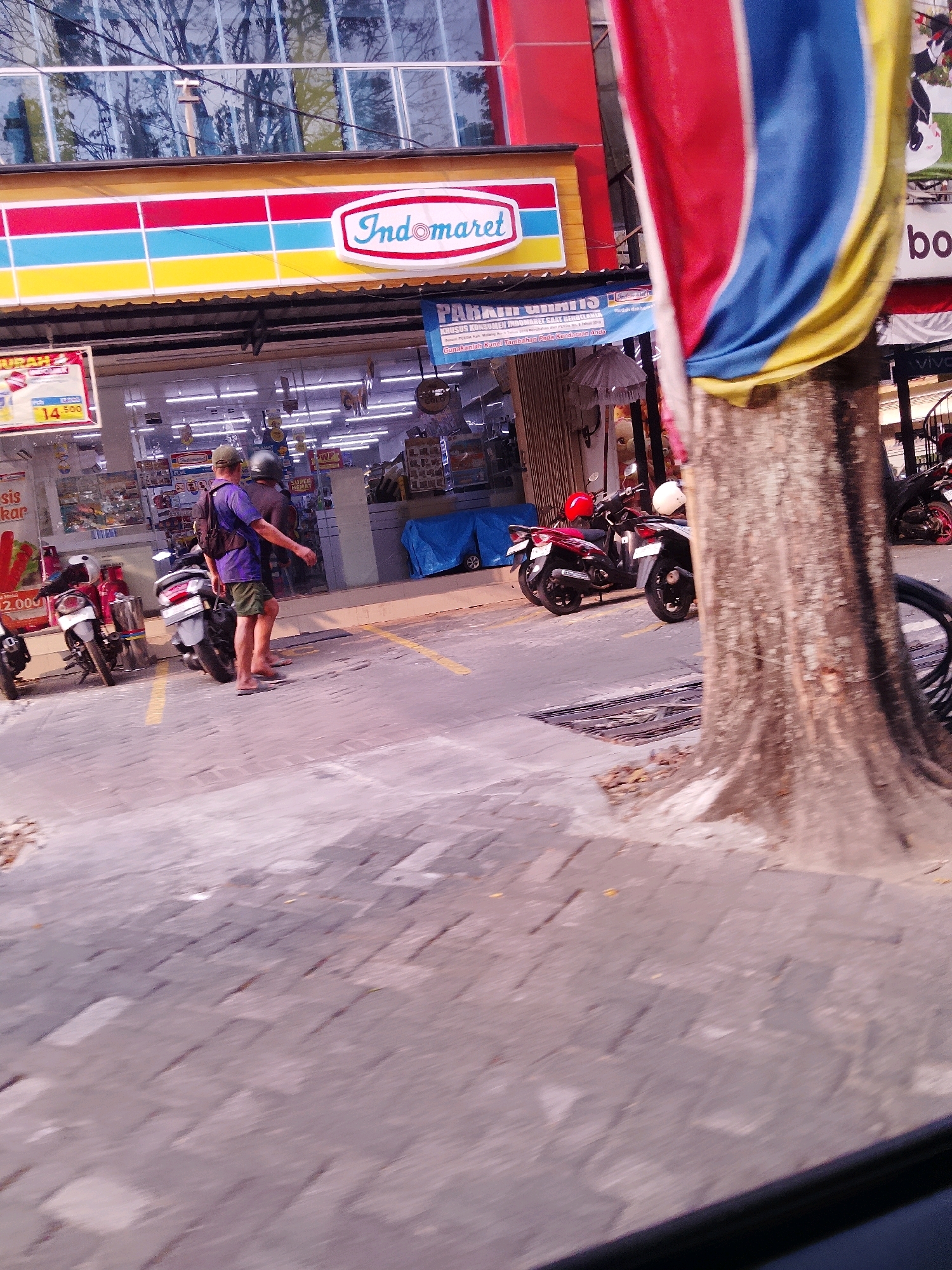 The image depicts a street scene in front of an Indomaret convenience store, likely in Malang, Indonesia. A man wearing a purple t-shirt, green shorts, a cap, and carrying a backpack is walking away from the camera. He is barefoot. Several motorcycles are parked on the sidewalk and street. The Indomaret store has a prominent sign with the brand logo. A large tree trunk is visible in the foreground, obscuring part of the scene. A flag with red, blue, and yellow stripes is also draped on the tree. The ground appears to be paved with cobblestones and asphalt. The overall lighting suggests it is daytime.