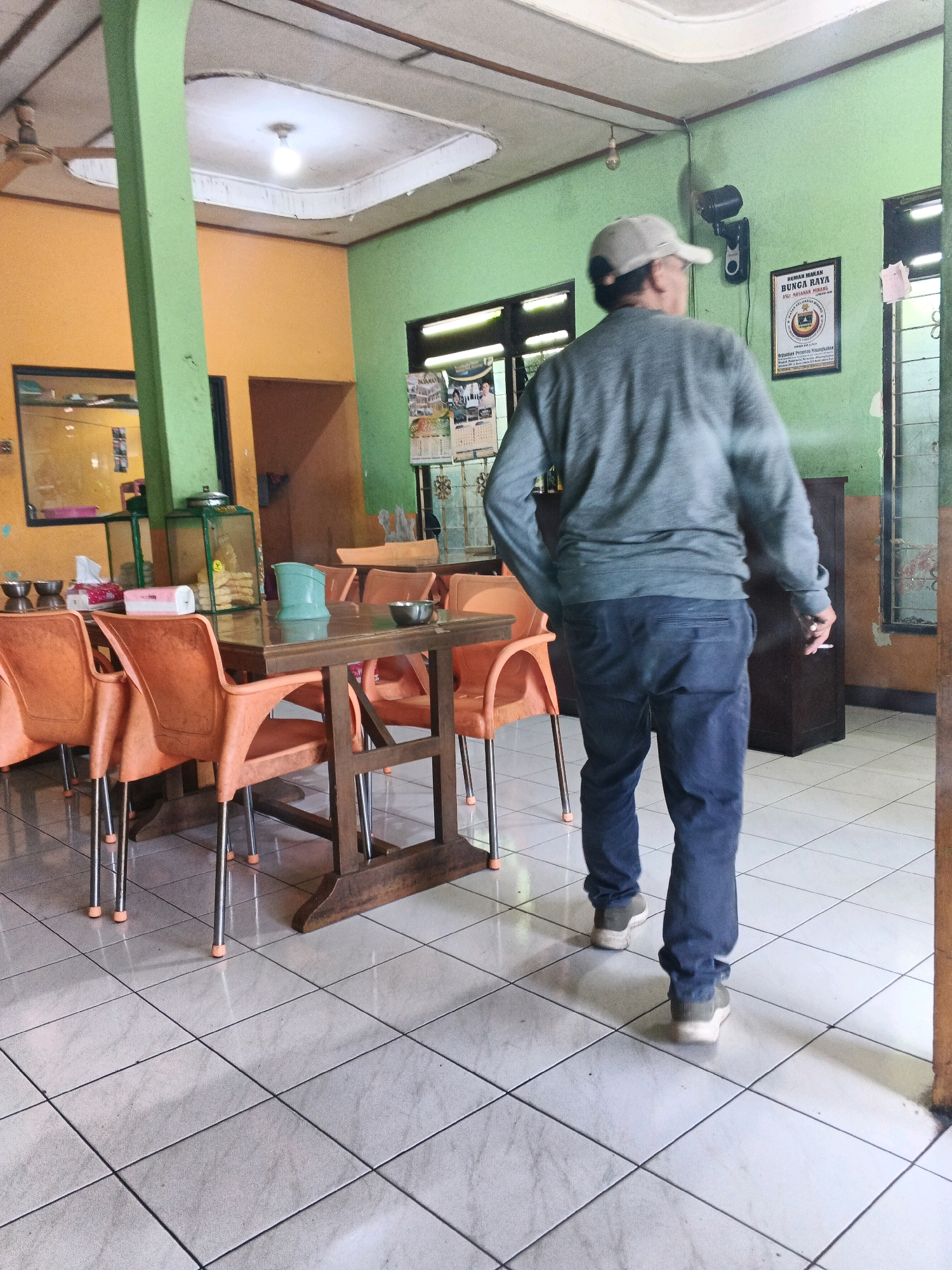 A man wearing a gray sweater, dark blue pants, a light-colored baseball cap, and sneakers walks away from the camera inside a brightly lit dining area. The man holds a cigarette in his right hand. The dining area has orange plastic chairs and wooden tables with brown wood legs. The floor is tiled with light-colored square tiles. The walls are painted green and orange. A ceiling fan is visible in the upper left corner. A framed certificate hangs on the green wall. The scene appears to be a restaurant in Ungaran, Indonesia, a city known for its culinary scene.