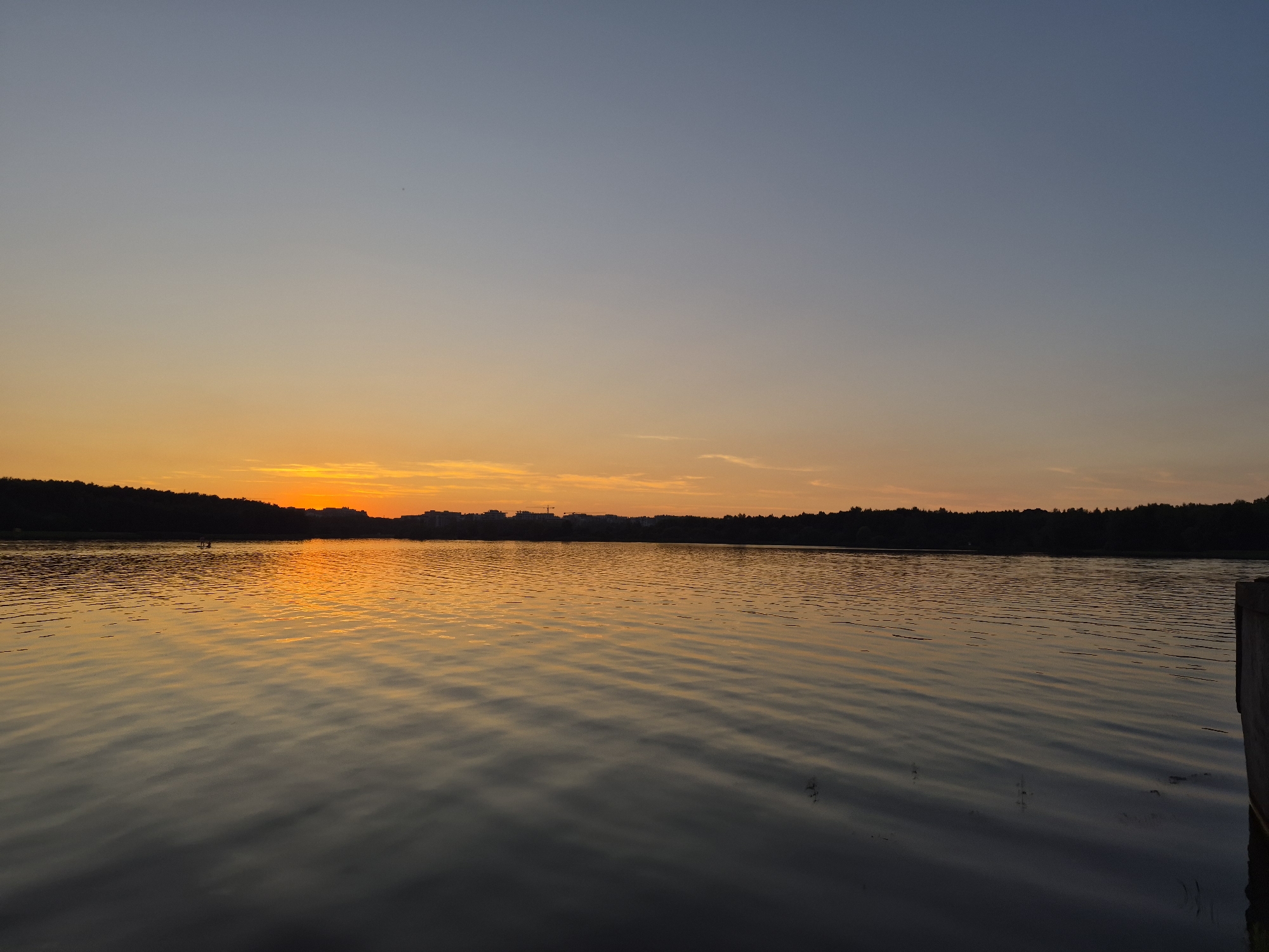 The image depicts a serene sunset over a body of water, likely a lake or large river, with a silhouetted tree line forming the horizon. The sky transitions from a clear, pale blue at the top to a vibrant orange and yellow gradient near the horizon, indicating the sun is setting or has just set. The water reflects the colors of the sky, creating shimmering ripples. In the distance, along the tree line, are faint outlines of buildings and possibly a construction crane, suggesting a developed area bordering the natural landscape. The foreground shows a dark, wooden structure on the right side of the frame, possibly a dock or pier. There are no visible people or animals in the image. The scene evokes a peaceful and tranquil atmosphere.