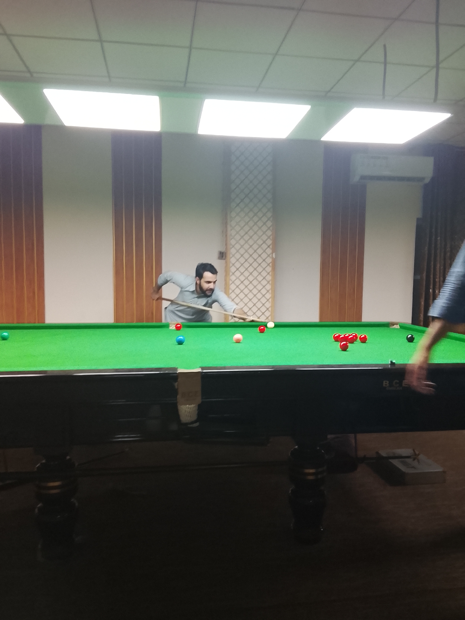 An indoor, eye-level shot captures a man playing snooker on a green-felted table. The man, with a beard and wearing a light-colored shirt, is leaning over the table, holding a cue stick, and aiming intently at the snooker balls. His focus is evident as he prepares to strike.
The snooker table is prominently featured in the foreground, with its vibrant green felt surface contrasted by a dark frame and decorative, dark wooden legs. Various snooker balls are visible on the table: a cluster of red balls, a black ball near a pocket, a pink ball, a blue ball, a green ball, a yellow ball, and the white cue ball. The text "B C E ENGLAND" is visible on the side of the table's frame.
The room's interior features a drop ceiling with white square ceiling tiles and several bright, rectangular overhead fluorescent light panels that provide ample illumination, creating some glare on the table's surface. The walls are paneled with light-colored sections interspersed with vertical dark wooden or striped inserts. An air conditioning unit is mounted on the wall in the background. A partially visible arm and hand of another person are seen on the far right of the frame, suggesting the presence of at least one other individual. The floor is carpeted in a dark color. The scene depicts an active game in what appears to be a billiards hall or game room.