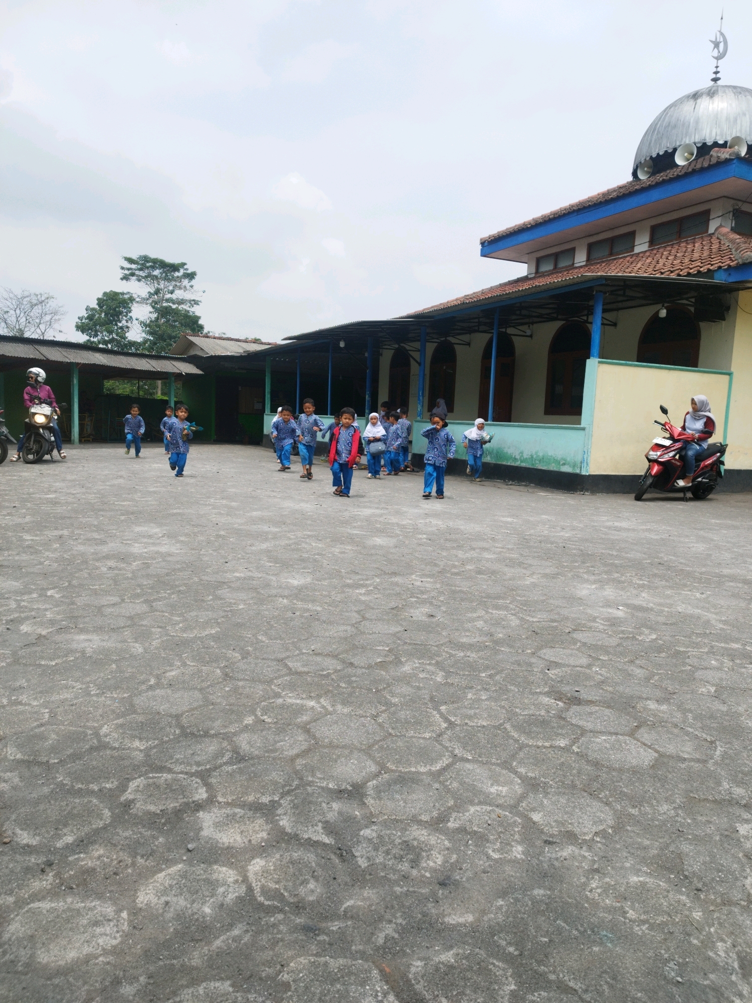 The image depicts an outdoor scene in front of a building with a mosque dome. Several children, dressed in blue and white uniforms, are running across a paved courtyard. Some are looking towards the camera. A woman wearing a headscarf is sitting on a red motorcycle parked on the right side of the image. Another person on a motorcycle is visible on the left side. The building appears to be a school or religious institution, with a mosque dome on its roof. The sky is overcast. The courtyard is paved with hexagonal stones. The environment suggests it is a warm climate, likely in Indonesia given the architecture and attire. The location is specified as Hargobinangun, Indonesia.