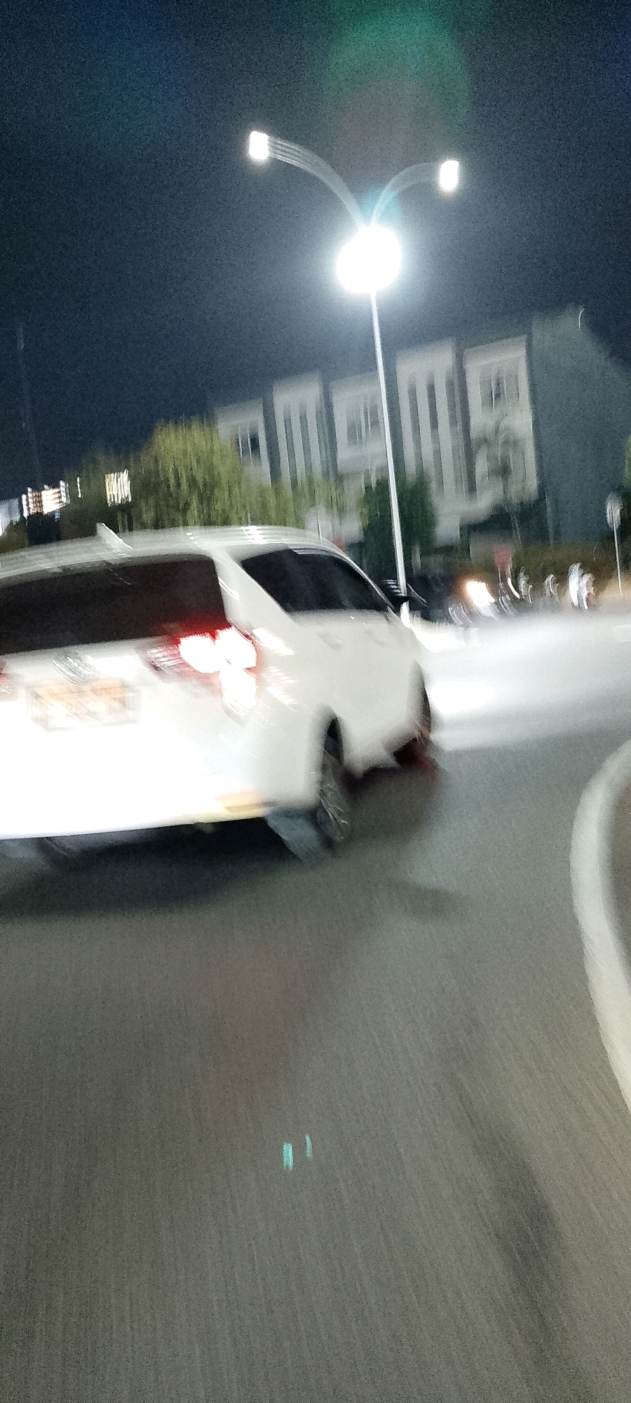 A white minivan is captured in motion at night on a road in Gunung Putri, Indonesia. The vehicle is angled towards the right of the frame, with its taillights illuminated and blurred due to the movement and low light conditions. In the background, there are multi-story buildings with prominent white architectural elements, possibly apartment complexes or offices. A street light with a dual-head design illuminates the scene, casting a bright glow and creating lens flare. Trees with green foliage are visible between the road and the buildings. The road surface appears to be asphalt, with a light-colored lane marker visible on the right side. The overall image is characterized by motion blur, suggesting a photograph taken while the vehicle was moving or from a moving platform.