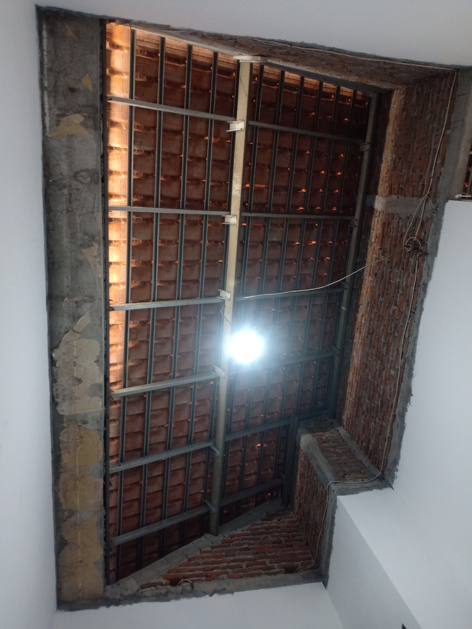 The image depicts the underside of a roof structure under construction, possibly a skylight or an open ceiling area. Reddish-brown tiles are visible, supported by a metal framework. The walls surrounding this area are made of rough concrete on the left and exposed brick on the right. A bright light source, likely a bulb, is illuminating the scene from within the roof structure, causing a glare. Electrical wiring is visible, running along the concrete and brick sections. The setting appears to be an indoor construction site. Based on the provided information, this scene is located in Semarang, Indonesia.