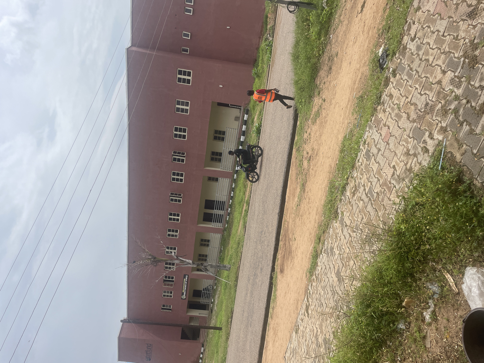 In Lokoja, Nigeria, a scene unfolds on a street in front of a large, maroon-colored building. The building, possibly an administrative or educational facility, features a repeating pattern of square windows. A sign on the building reads "DIRECTORATE OF SIWES" and "TETFUND."
Two individuals are present on the asphalt road. One, on a motorcycle, is seen from the side, wearing dark clothing. The other individual, a person wearing a bright orange safety vest over a red shirt and dark trousers, walks away from the motorcycle, facing right. They appear to be a pedestrian.
The environment includes a paved walkway with interlocking brick pavers bordering a patch of green grass and dirt. Overhead, power lines crisscross against a cloudy sky. The overall impression is one of an ordinary day in a functional, possibly institutional, setting.