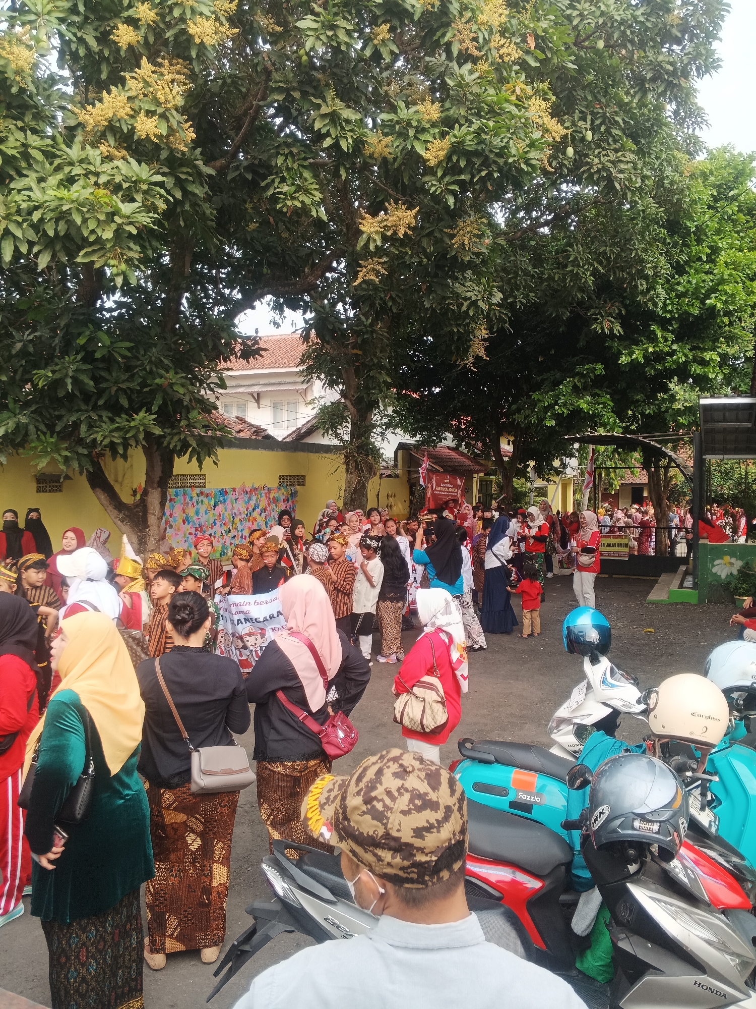 In the city of Sokanegara, Indonesia, a group of people, predominantly children, are gathered outdoors. Many of the children are dressed in traditional Indonesian attire, including batik sarongs and distinctive head coverings. Some are holding a banner that reads "main bersama demi bangsa dan negara." This suggests a cultural event or celebration, possibly a parade or school activity. Adults are also present, some supervising the children and others observing the proceedings. The environment appears to be a street or schoolyard, with trees providing shade. Several motorcycles are parked in the foreground, indicating this is likely a public gathering. The overall atmosphere seems festive and communal.