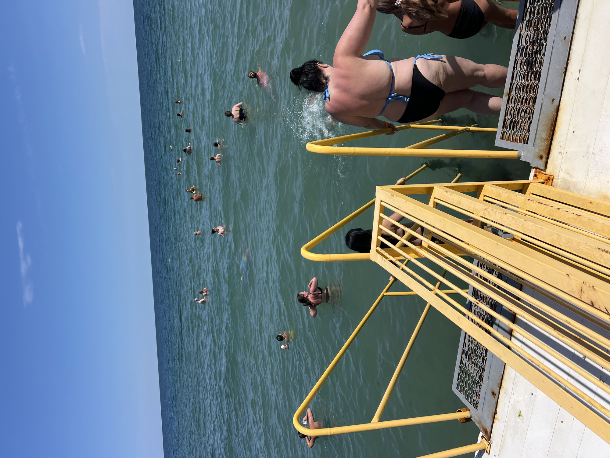 A group of people are swimming in the Black Sea off the coast of Odesa, Ukraine. The water is a greenish-blue and appears calm. In the foreground, a yellow metal stairway leads down into the water, with some people on the steps. Further out in the water, many other individuals are visible swimming or wading. The sky is clear and blue, with a few wispy clouds. The scene depicts a typical summer day at a beach or pier.