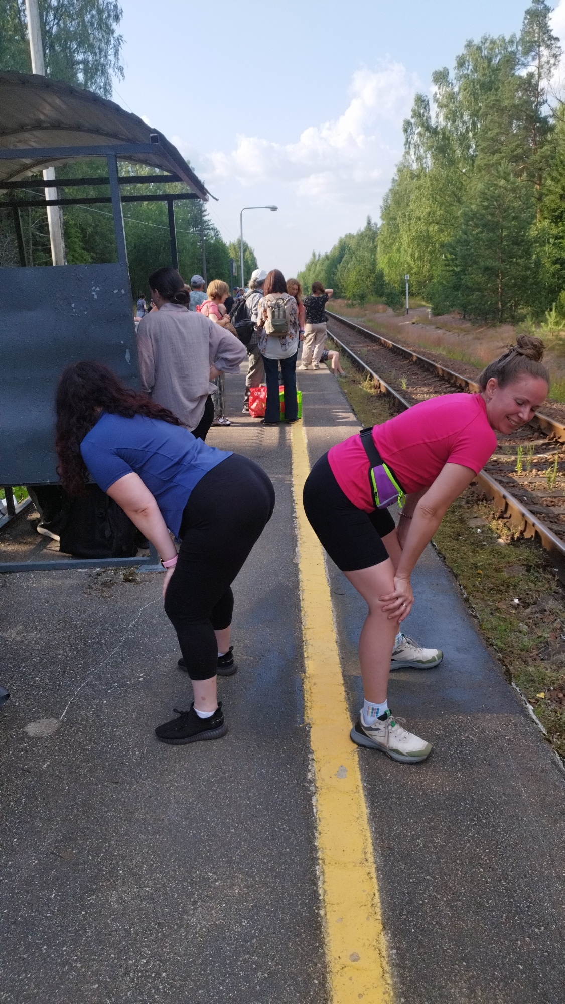 Two women are standing on a train platform, bending forward with their hands on their knees. They are both wearing athletic attire. The woman on the left has long, curly dark hair and is wearing a blue shirt and black leggings. The woman on the right has her hair pulled back in a bun and is wearing a pink shirt, black shorts, and athletic shoes. Other people are visible on the platform in the background, some are standing and others appear to be sitting or lying down. The platform is paved and has a yellow line marking the edge. Train tracks run alongside the platform, and trees and foliage are visible in the background. The sky is partly cloudy. The location is identified as Tarasovskoe, Russia, specifically on a train platform.