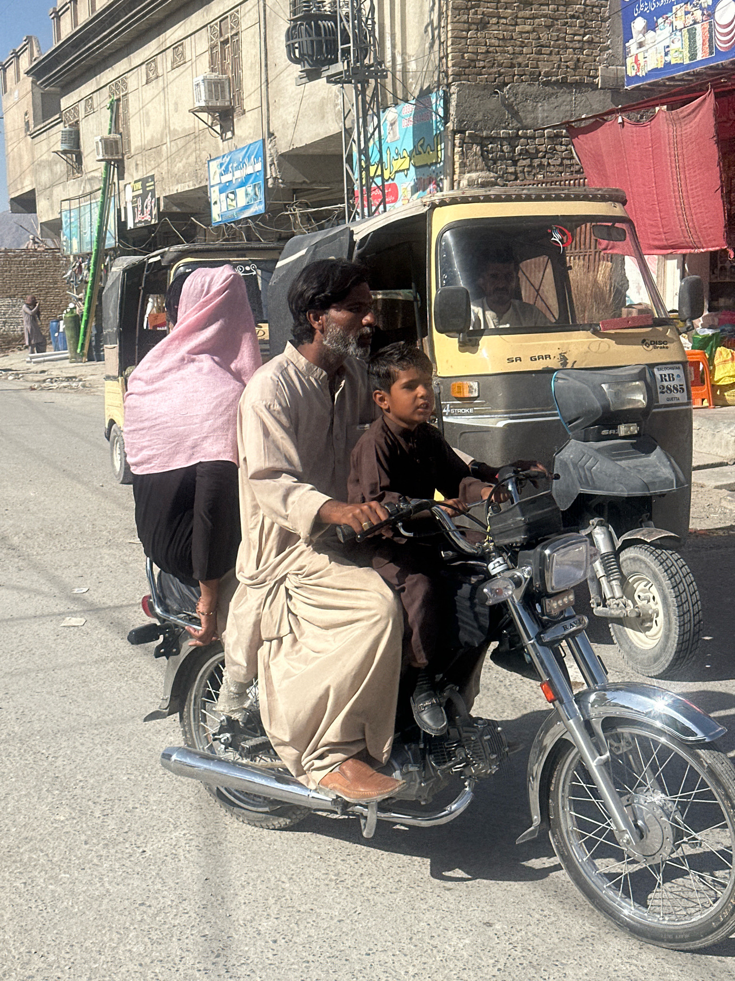 In Quetta City Tehsil, Pakistan, a street scene unfolds with a man and a young boy on a motorcycle, positioned in the foreground. The man, with a beard, is seated behind the boy and appears to be operating the motorcycle. He is wearing a light brown, loose-fitting traditional garment. The boy, in a dark brown outfit, is in front, holding the handlebars. Behind them, a woman draped in a bright pink headscarf and black clothing is seated on the back of the motorcycle. The motorcycle is a modern style with chrome accents.
To the right of the motorcycle, a yellow three-wheeled auto-rickshaw is visible. The auto-rickshaw has a license plate that reads "RB 2885 QUETTA". The driver of the auto-rickshaw is partially visible through the windshield. In the background, buildings with a rustic, somewhat aged appearance line the street, with signs and wires adding to the urban environment. A red fabric hangs from one of the buildings on the right. The ground is paved, and there are some scattered papers visible on the road. The overall atmosphere suggests a typical day in a bustling urban area of Pakistan.