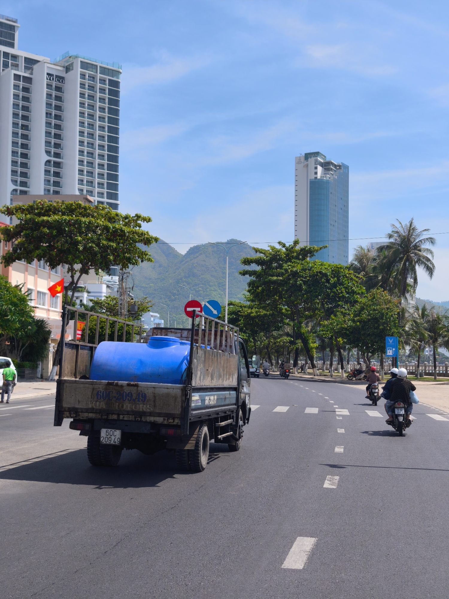 In a city scene, a dark-colored truck transports a large blue plastic barrel on its flatbed. The truck is positioned on the right side of a multi-lane asphalt road. The barrel is oriented horizontally and secured within the truck's cargo area. The truck's license plate reads "60C 209.49".
To the left of the truck, a white car is parked near the curb. A person wearing a green shirt is walking away from the camera on the sidewalk. Adjacent to the sidewalk, a vibrant green tree with red accents on its leaves stands prominently. Further down the street, the Vietnamese flag is visible, attached to a pole.
The background features a row of tall, modern buildings. The building closest to the viewer is white with dark-framed windows. Another taller, slender building with a blue glass facade is visible further in the distance. Lush green trees line the street, and a mountainous landscape forms the backdrop under a clear blue sky with sparse white clouds.
On the road, several motorcycles are in motion. Two individuals on a motorcycle are traveling ahead of the truck, while other motorcycles are seen further down the street. Traffic signs, including a red and white "no entry" sign and a blue directional arrow, are posted on the right side of the road. The scene captures a slice of urban life, likely in a coastal city given the presence of palm trees and the expansive sky.