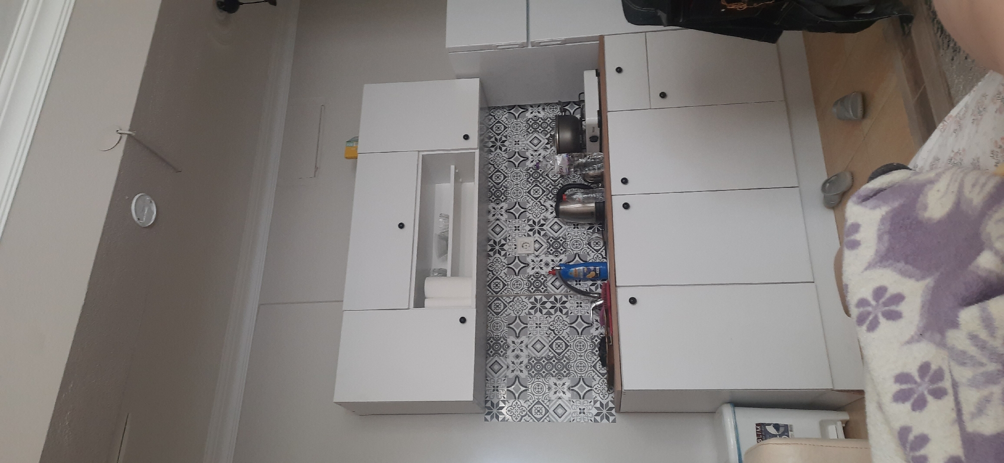 The image depicts the interior of what appears to be a modern kitchen or kitchenette. The primary features include a series of white cabinets and drawers, some of which are open. The open drawer reveals a patterned backsplash and kitchen essentials such as kettles and cleaning supplies. The floor is tiled, and there are a few pairs of flip-flops or slippers scattered around. A partially visible blanket with floral patterns suggests a residential setting.
Without additional information, pinpointing the exact location in Kemer, Turkey is not possible. Kemer is a popular tourist destination, and the decor suggests a possible rental apartment or hotel room.
