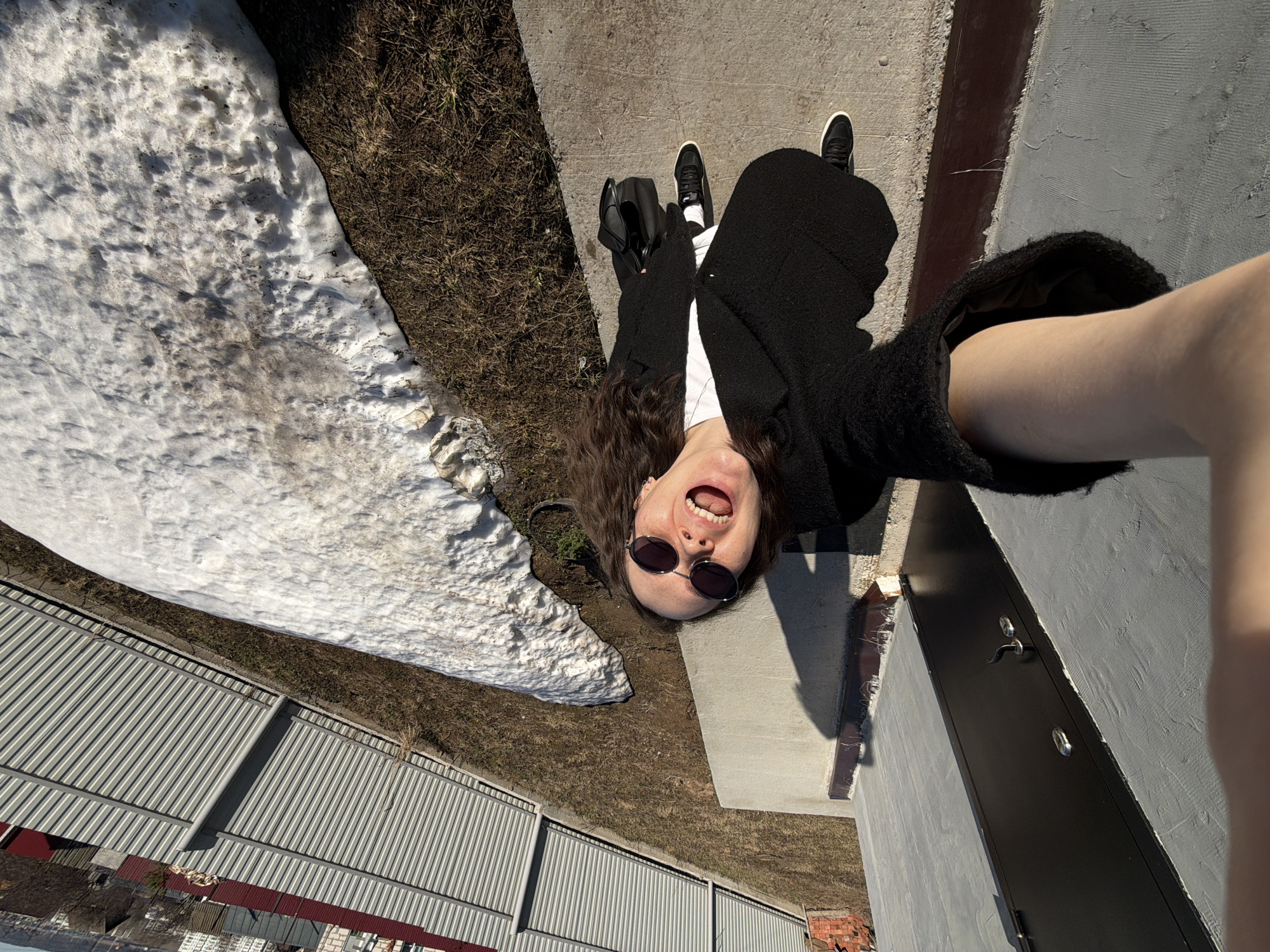 A medium shot shows a woman lying on her side on a concrete surface, holding up a smartphone. She has long, curly brown hair and wears black sunglasses, a white shirt, and a black jacket. Her mouth is open in a wide smile or shout. Her legs are extended, and she wears black and white sneakers. Next to her is a black purse.
To the left of the woman is a patch of melting snow and a grassy area. In the background, there is a metal fence, some red brick structures, and what appears to be a city skyline. To the right of the woman is a gray wall with a dark gray door. The photograph appears to have been taken in Chaikovsky, Russia.