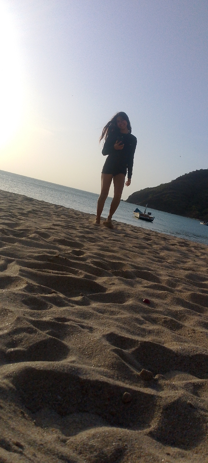 A low-angle photograph depicts a beach scene, likely taken during late afternoon or early morning based on the lighting. The foreground is dominated by uneven, light brown sand with distinct footprints and small indentations. A single small, dark red, irregularly shaped object rests on the sand in the lower right quadrant.
In the midground, a female individual stands barefoot, angled slightly to the viewer's right. She has long, dark hair, appears to be smiling, and is wearing a long-sleeved dark top and dark shorts. Her right arm is bent, and she is holding a dark-colored smartphone-like device to her chest, possibly engaging with it or capturing the image. Her left arm is slightly bent at her side. Her bare feet are partially obscured by sand, suggesting she is standing still or moving slowly.
Behind the individual, the calm, light blue-green ocean extends horizontally across the frame. A small, light-colored fishing boat with a dark hull and a short mast is anchored in the water, positioned to the right of the person. Further in the background, a large, dark green, tree-covered hill rises from the water's edge, defining the coastline. The sky above is a clear, light blue, transitioning to a brighter white-yellow hue on the left, indicative of the sun's position. The overall scene suggests a coastal area within Juan Griego, Venezuela, consistent with its geography.