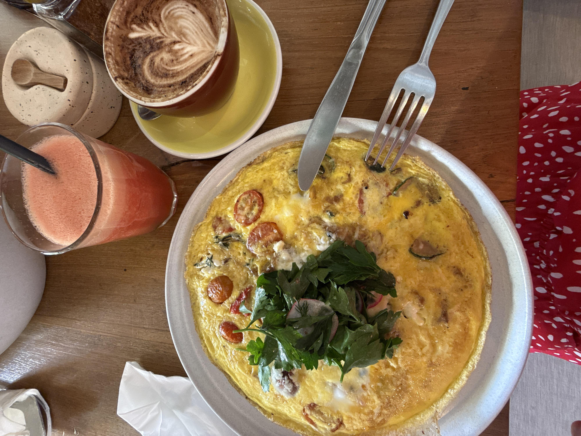A high-angle shot reveals a meal setting on a wooden table, featuring an omelet on a white plate, garnished with fresh greens and accompanied by a fork and knife. Beside the plate, a glass of pink smoothie stands next to a cup of coffee with latte art, set on a yellow saucer. A small white container with a wooden spoon rests near the coffee. A glimpse of a red and white patterned fabric is visible in the background. The scene suggests a cafe setting, possibly in North Kuta, Indonesia.