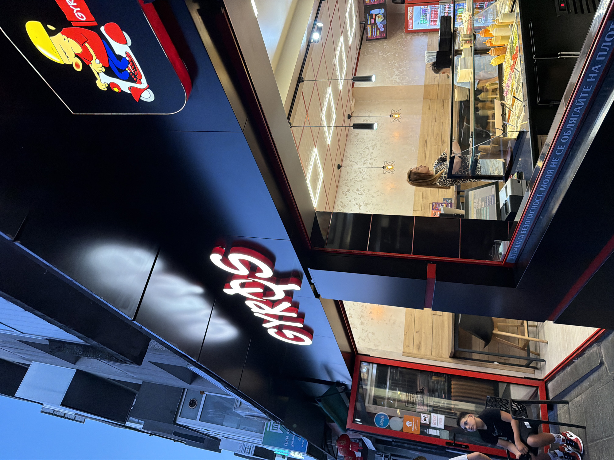 The image shows the exterior of a "GYROS" restaurant located in Burgas, Bulgaria. The restaurant has a prominent black facade with illuminated white lettering spelling out "GYROS". Above the entrance, a cartoon character on a scooter is depicted in a circular sign. The entrance is open, revealing the interior with a counter displaying various food ingredients. A young woman with shoulder-length brown hair, wearing a leopard print top, is standing behind the counter, appearing to serve customers. To the left of the entrance, a young male with dark hair, wearing a black t-shirt and shorts, is seated on a chair outside the establishment, looking upwards. The street is paved, and the building appears to be multi-story with architectural balconies. The sky is visible in the upper left corner, indicating it is either early morning or late evening. There are several signs and logos visible near the entrance, including those for Wolt and "Takeaway.com". A red Coca-Cola cooler is visible inside the restaurant to the right.