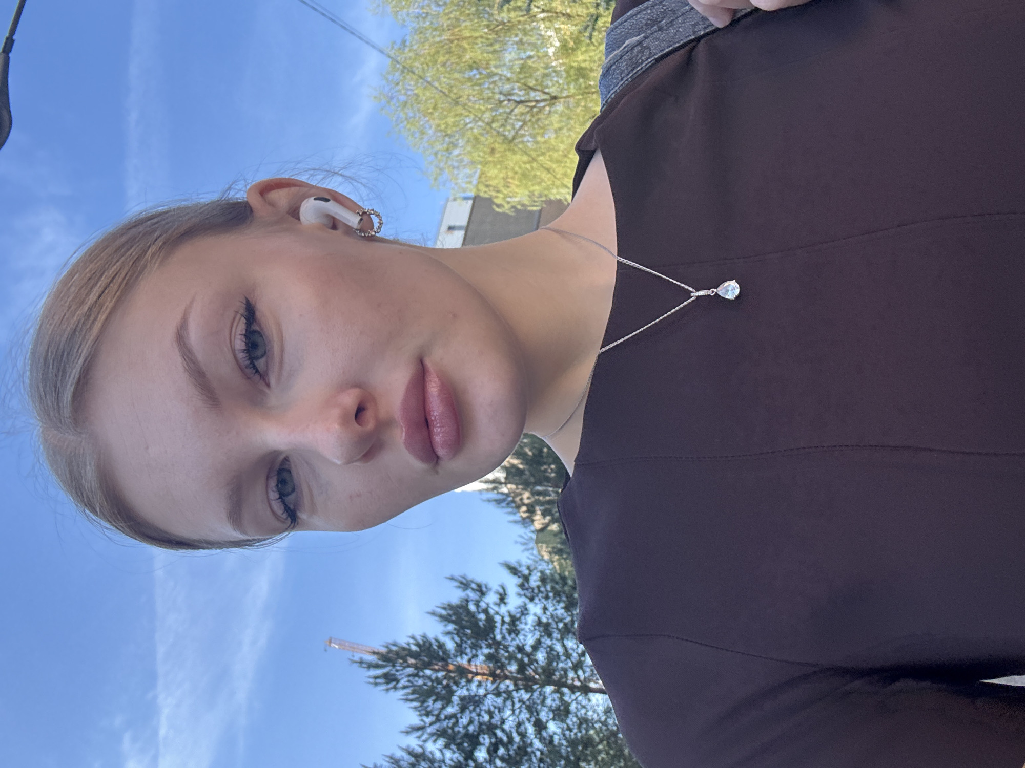 A medium shot depicts a Caucasian woman with light blonde hair, wearing a silver necklace with a teardrop pendant, and AirPods in her ear. The background features a blue sky, trees, and a building under construction with a crane visible. The photo may be taken in Perm, Russia, a location not definitively discernible from the visual content provided.