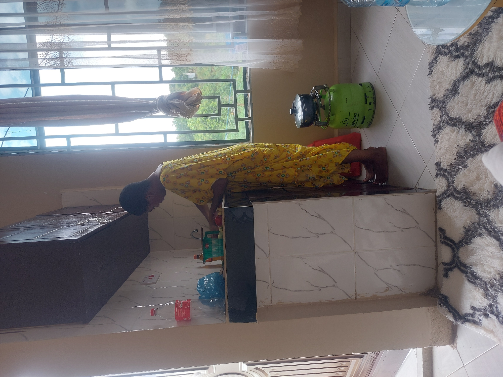 A woman in a yellow floral dress is leaning over a kitchen counter. On the counter is a faucet and some bottles. A dark cabinet is visible to the left of the counter. A green gas tank sits on a red pad next to the counter, near a window. The window looks out onto a green, somewhat rural, landscape. The floor is tiled, and a patterned rug is partially visible. The location is potentially Dar es-Salaam, Tanzania.
