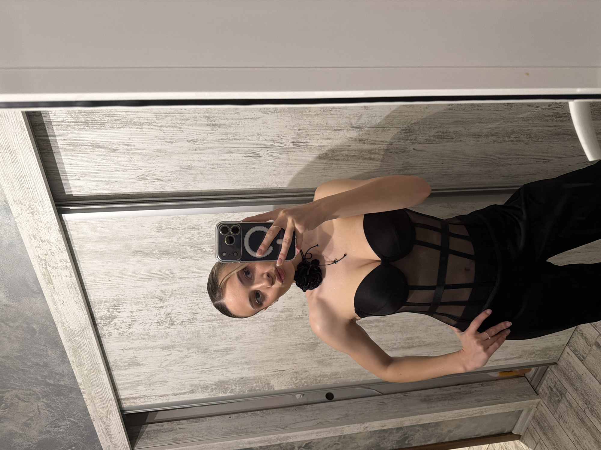 A young woman is taking a selfie in a mirror. She is wearing a black corset-style top and black pants. The location appears to be an indoor space with a wooden-look surface behind her and a mirror. The photograph does not contain enough information to determine the specific location in Plovdiv, Bulgaria.