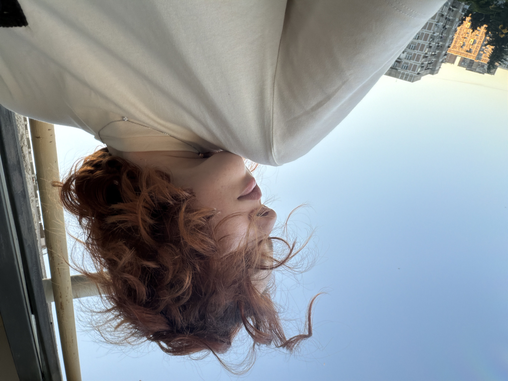 The image is an upside-down, low-angle shot of a fair-skinned person with auburn, curly hair. The subject is wearing a light-colored shirt and a thin silver necklace with a small pendant. The person's head is tilted back, exposing the sky as the background. In the distance, a building complex with a beige facade and multiple windows is visible, identifiable as architecture commonly found in Tbilisi, Georgia. A metal railing is visible in the bottom left corner, suggesting the subject is positioned on a balcony or elevated structure.