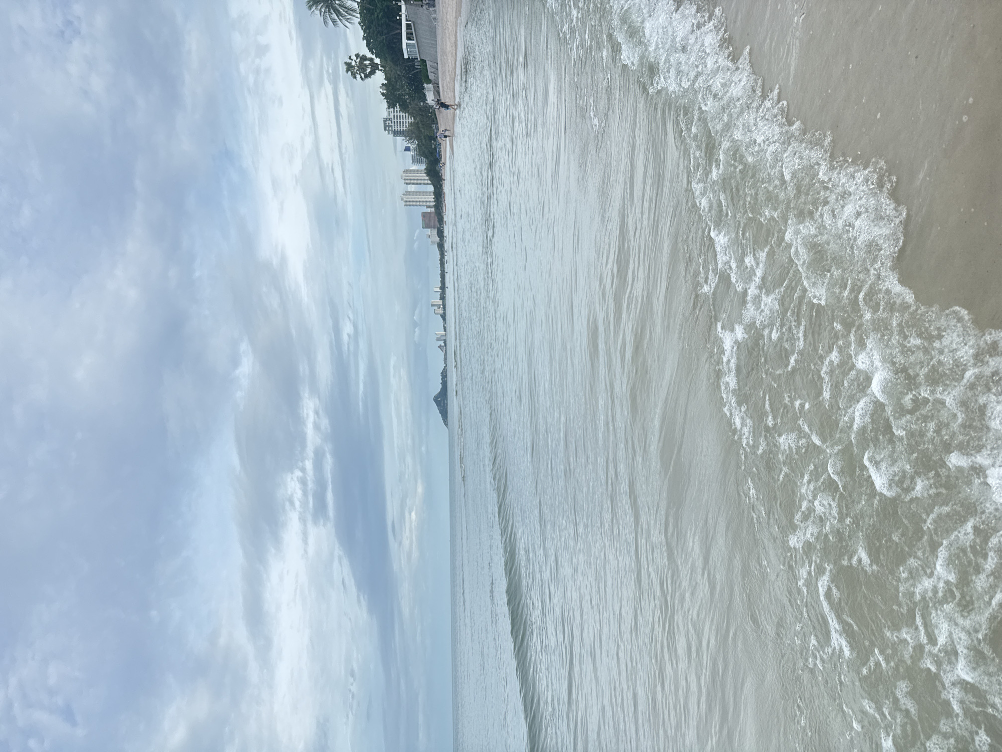 The image depicts a serene beach scene in Hua Hin, Thailand. The sky is overcast with soft, diffused light, creating a calm atmosphere. Gentle waves lap onto the sandy shore, their foam creating a delicate pattern as they recede. In the distance, a line of buildings, including several tall hotels and residences, marks the urban edge of the coastline. Palm trees and lush vegetation are visible along the treeline, adding a tropical element to the scenery. A few people can be seen strolling along the beach, some closer to the water's edge and others further up towards the buildings, suggesting a leisurely day. The overall impression is one of tranquility and a pleasant coastal environment.
