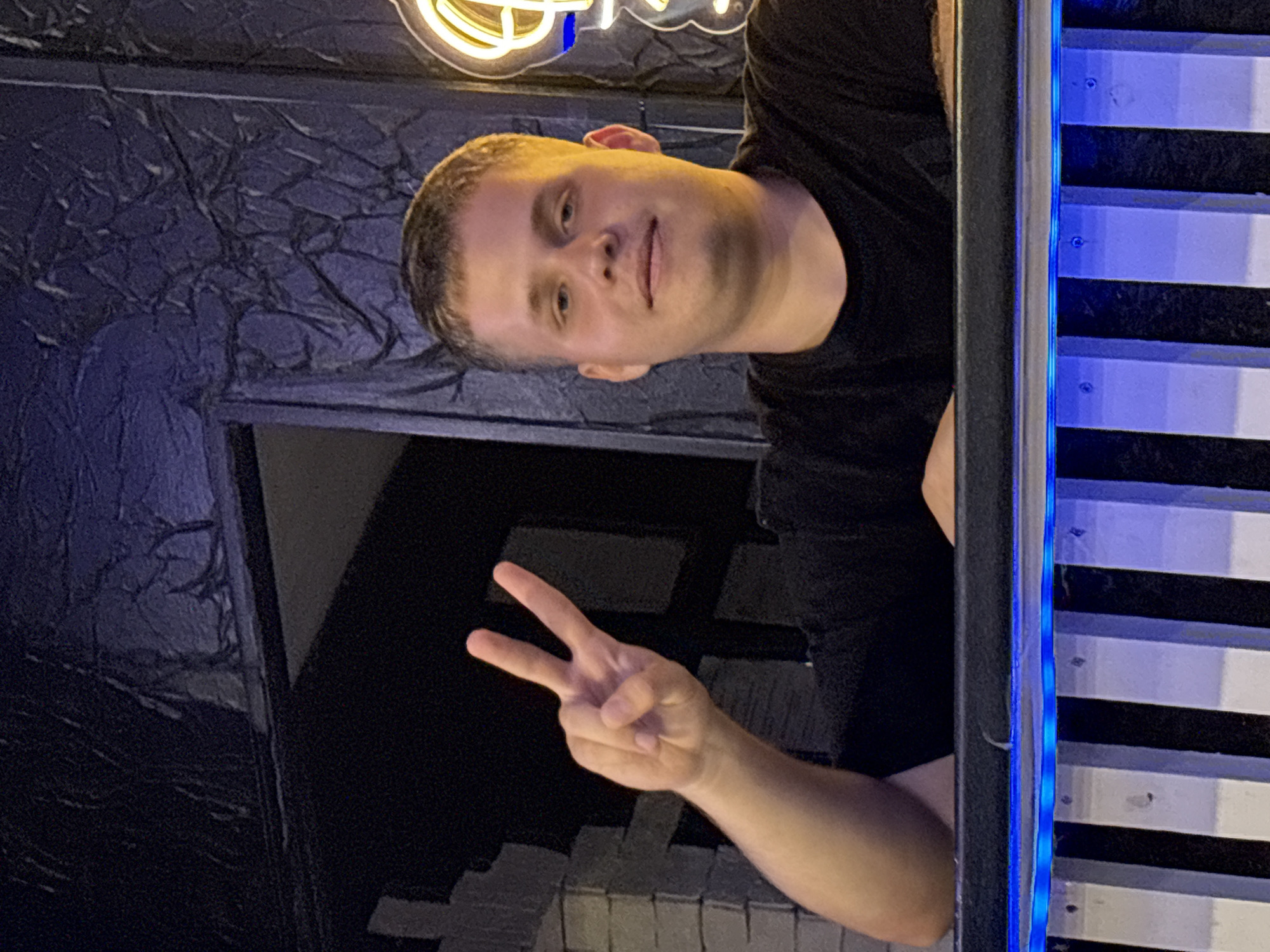 A medium shot shows a young Caucasian male with short blonde hair and blue eyes, wearing a black t-shirt. He is positioned behind a railing with blue LED lights. His right arm is raised, and he is making a peace sign with his index and middle fingers extended. The background is dark and textured, with hints of a dark, crinkled material resembling fabric or wallpaper. A neon light fixture is visible on the right side of the frame. The image appears to have been taken at night, possibly in an outdoor setting such as a bar or entertainment venue. The specific location in the city of Gorodets, Russia cannot be determined from the image alone.