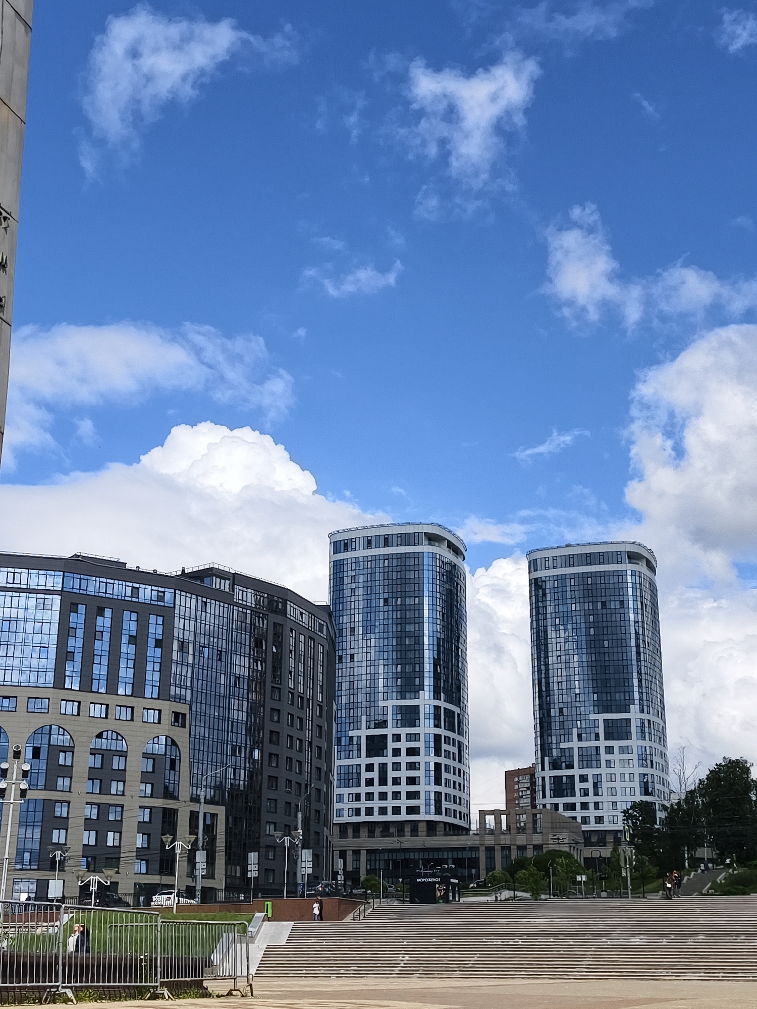 This image presents a vibrant urban scene in Izhevsk, Russia, under a bright blue sky filled with scattered white cumulus clouds. The foreground features a broad, light-colored concrete staircase with numerous steps, suggesting a public square or elevated pathway. A metal barrier runs along the left side of the stairs, enclosing a patch of green.
The mid-ground and background are dominated by modern architecture. On the left, a multi-story building displays dark, reflective glass facades interspersed with sections of lighter, possibly beige, material featuring distinctive arched windows on its lower levels. To its right stand two prominent, cylindrical skyscrapers. These tall buildings are characterized by their dark glass exteriors highlighted with intricate white or light grey geometric patterns, giving them a sleek, contemporary appearance. Behind them, other buildings and structures, including one with a reddish-brown hue, are partially visible.
Several people are present in the scene, walking or standing in different areas. One person is visible near the metal fence on the left, and a couple of individuals can be seen further back near the base of the two cylindrical towers on the right. Streetlights and various poles are also visible throughout the urban landscape.
A notable detail is a small kiosk-like structure situated at the base of the buildings on the right, which displays the Russian word "МОРОЖЕНОЕ," indicating an "ICE CREAM" vendor. The time of day appears to be sunny daytime, likely morning or early afternoon, given the clear skies and soft lighting. The overall impression is one of a bustling, modern city on a clear and pleasant day.
