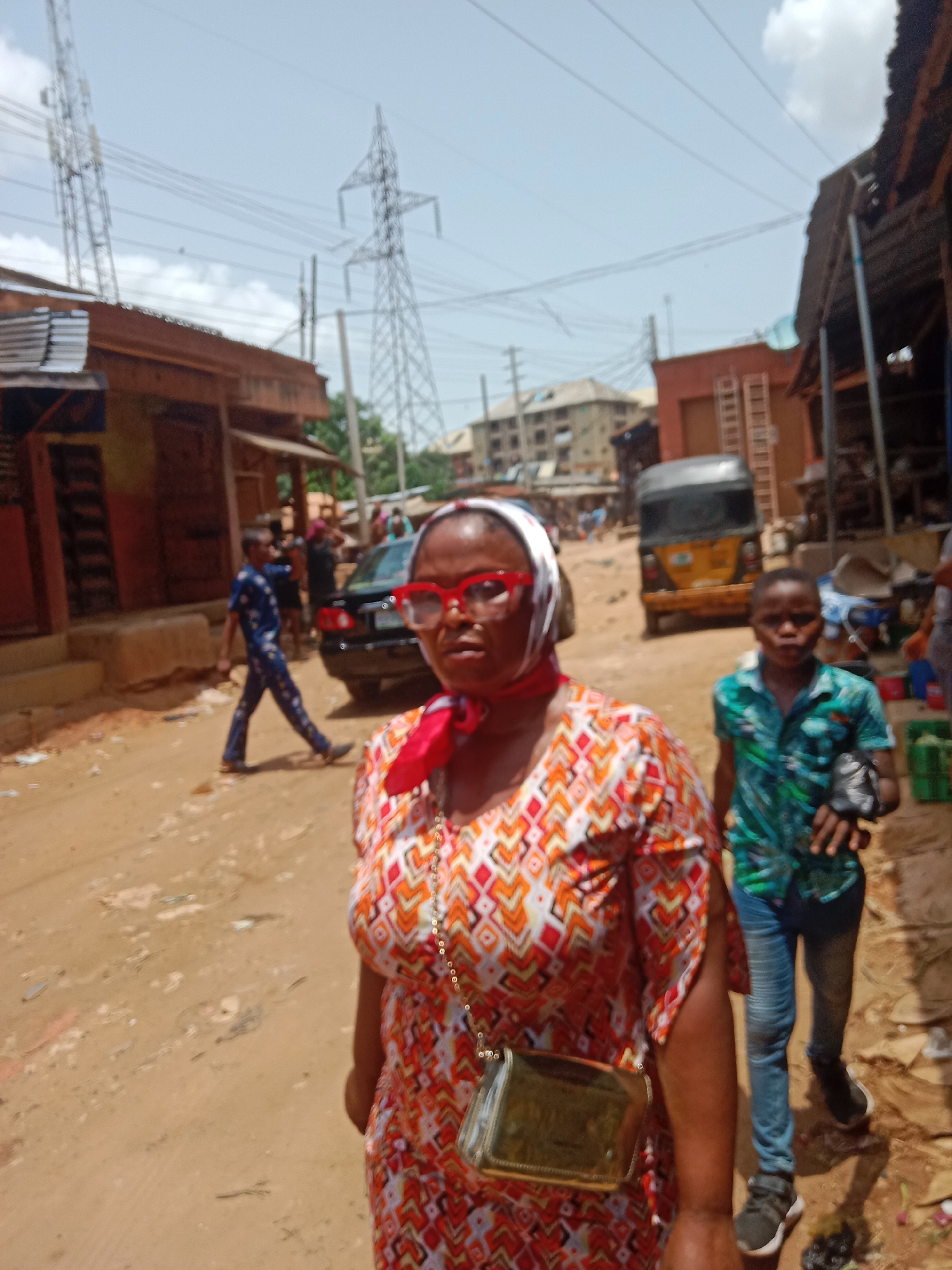 A woman wearing a patterned dress, red glasses, and a headscarf is in the foreground of a street scene in Onitsha, Nigeria. A young boy in a green shirt and jeans walks beside her. Behind them, a yellow auto-rickshaw is parked on the side of the road. Other people and vehicles are visible further down the street. The environment consists of various shops and buildings, with overhead power lines visible in the sky. The ground is a dirt road.