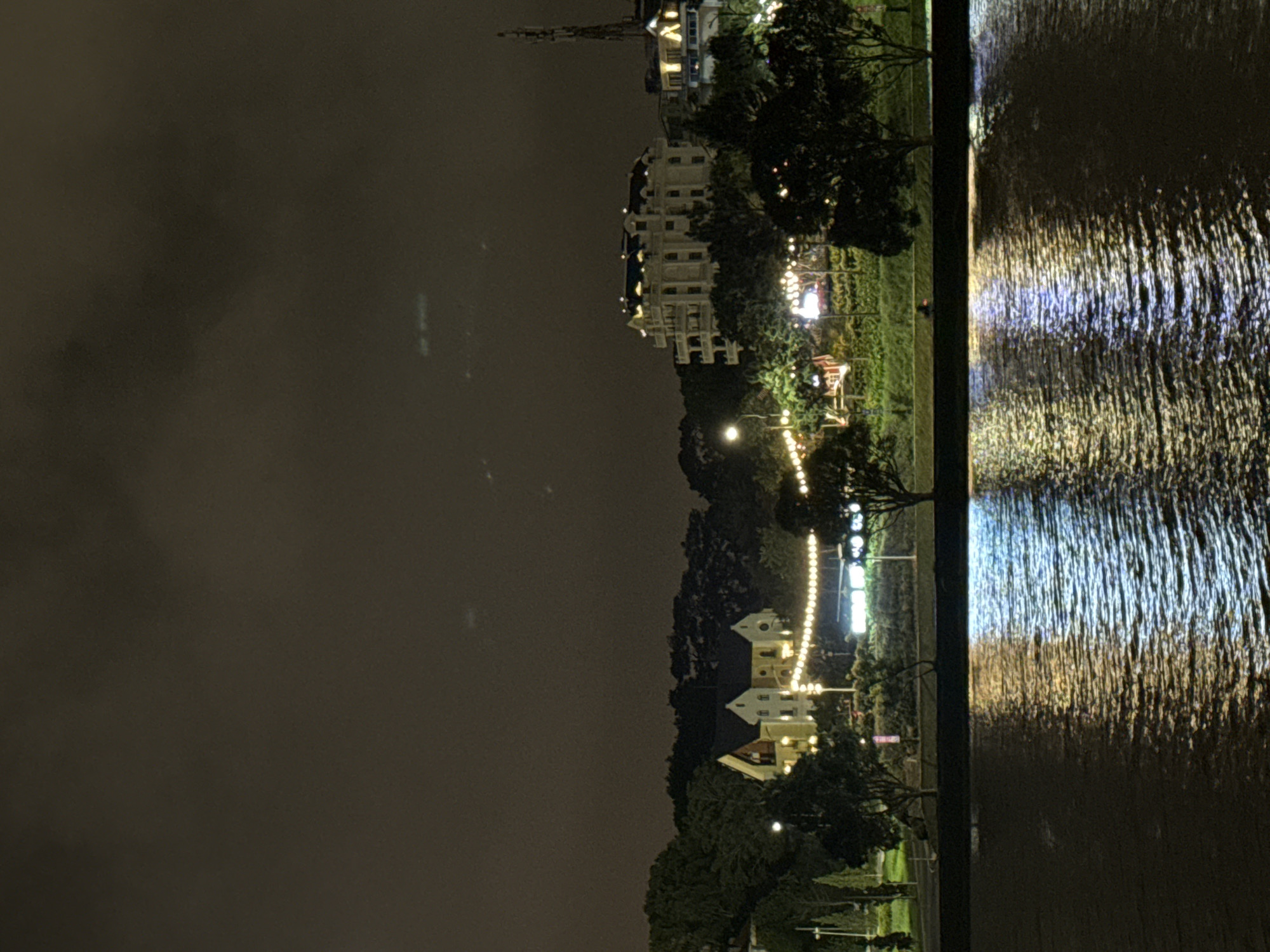 The image presents a serene nighttime view of a townscape alongside a body of water, set in Da Lat, Vietnam. The scene is illuminated by numerous artificial lights, creating a tranquil yet vibrant atmosphere.
The upper portion of the frame is dominated by a dark, cloudy night sky, offering a muted backdrop to the illuminated scene below.
On the right side, a dark body of water, likely a lake or river, reflects a multitude of lights from the opposite bank. These reflections appear as elongated, shimmering streaks of warm yellow, bright white, and occasional blue hues, indicating a gently rippled surface. A dark, vertical structure, possibly a quay or embankment, borders the water on the left, separating it from the land.
The left side of the image showcases the town's landscape. Several multi-story buildings, primarily light-colored with illuminated windows, stand amidst lush green vegetation. One building, towards the top left, features a distinctive pointed spire, hinting at European-influenced architecture common in Da Lat. The area is richly lit by various sources: numerous strings of warm-toned fairy lights are draped across trees and along pathways, contributing to a festive and inviting ambiance. Brighter streetlights and lights emanating from within the buildings further illuminate the scene. Small, decorative structures, including one with a red roof, are discernible within the verdant landscape.
No specific activities or people are visible, emphasizing the peaceful nature of the evening. The overall impression is one of a charming and picturesque nocturnal setting, typical of a popular leisure or tourist destination. No visible text is present in the image.