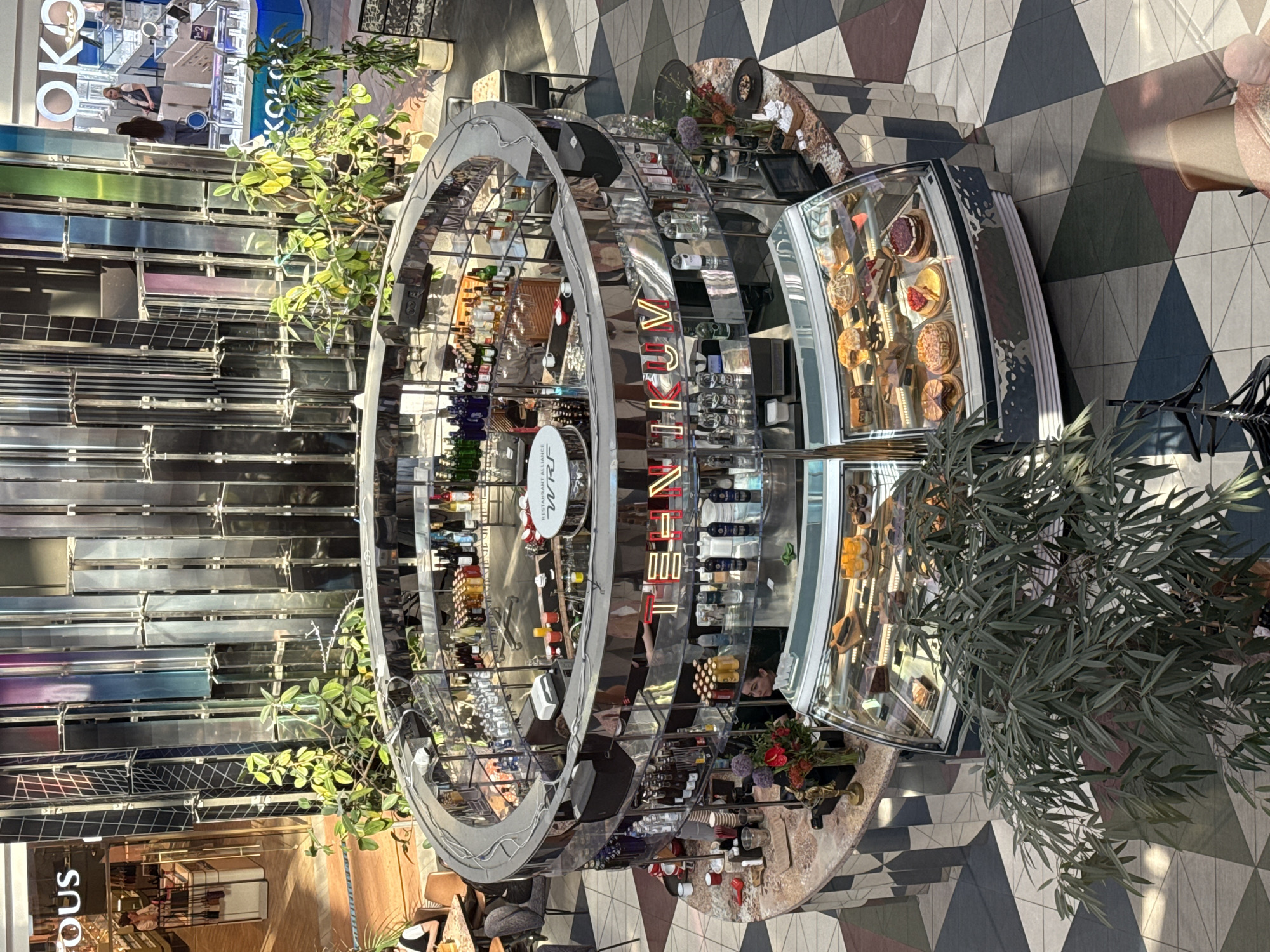 The image shows an indoor view of a modern retail space in Moscow, Russia, featuring a circular bar or food service area. The structure has a metallic, mirrored frame, with shelves displaying bottles of wine and other alcoholic beverages. One section of the structure includes a refrigerated display case containing pastries, cakes, and other desserts. The words "RESTAURANT ALLIANCE" are visible on a sign within the circular structure. The surrounding floor is tiled with a geometric pattern of triangles in various colors. Plants in large pots are strategically placed around the structure. The retail space appears to be part of a larger shopping mall or commercial area, with other stores visible in the background. There are no identifiable people in the image.