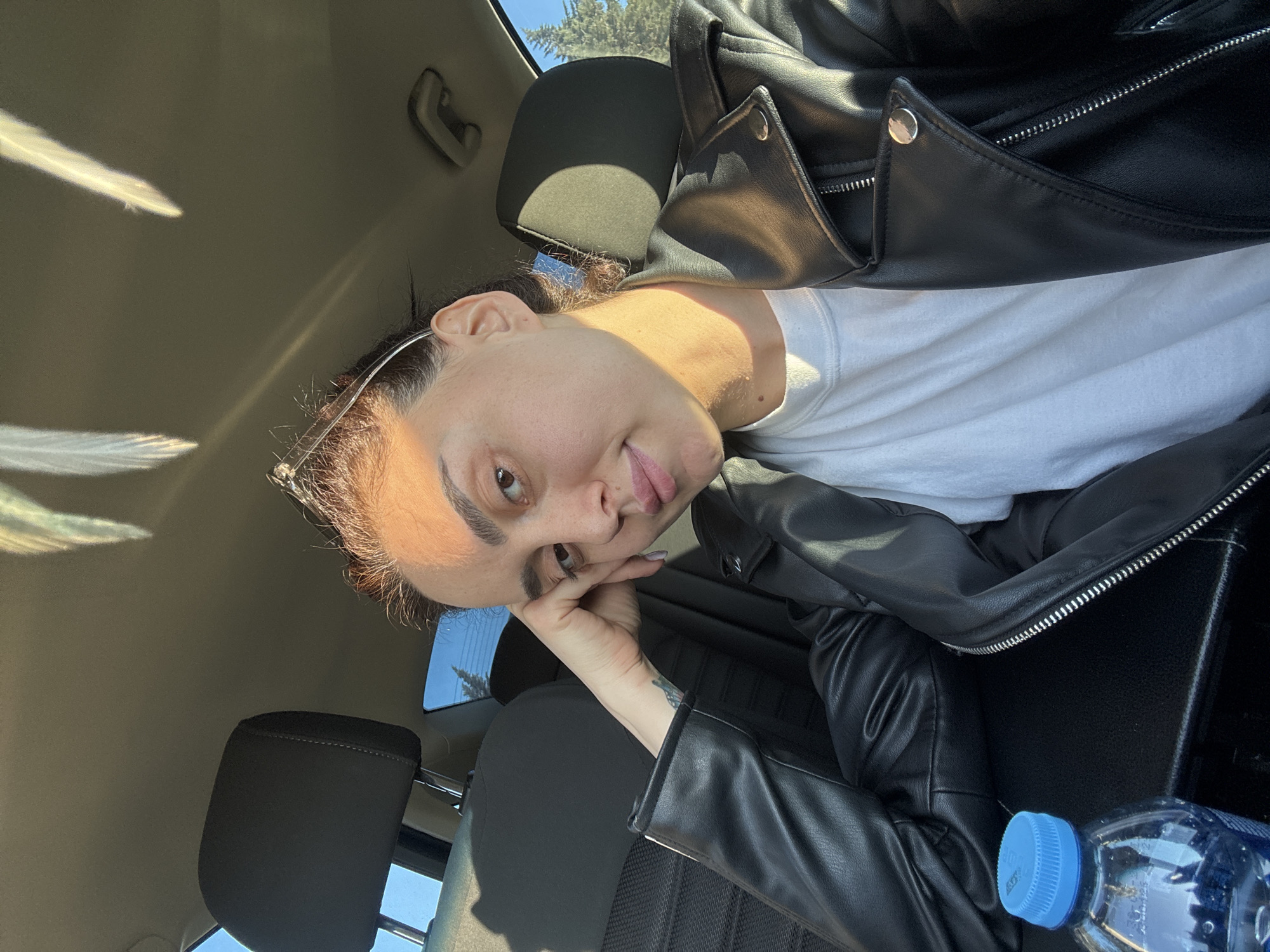 The image appears to be taken inside a car. A person is sitting in the driver's seat, wearing a black leather jacket and a white t-shirt. Their head is resting on their hand, and they are looking directly at the camera with a slight smile. Sunglasses are perched on their head. A bottle of water is visible in the bottom right corner of the frame. The interior of the car is visible, including the seats and the dashboard. The location appears to be in Tbilisi, Georgia, based on visible cues from the surroundings.