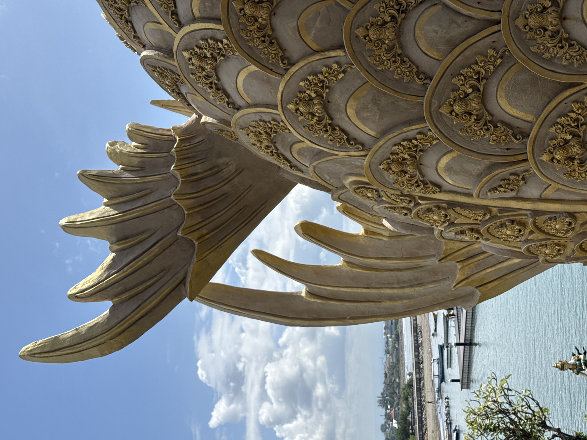 A low-angle, color photograph depicts a portion of a golden-colored sculptural representation of a dragon, seemingly part of a larger statue, against a partially cloudy sky, with a glimpse of a cityscape and harbor in the background. The dragon's scales are represented by overlapping circular patterns adorned with floral-like motifs, and it appears to have a wing or fin composed of multiple curved sections. The background reveals a body of water, likely a harbor, with docks and boats, and a cityscape in the distance with buildings and vegetation. The location is consistent with the harbor front in Denpasar, Indonesia.